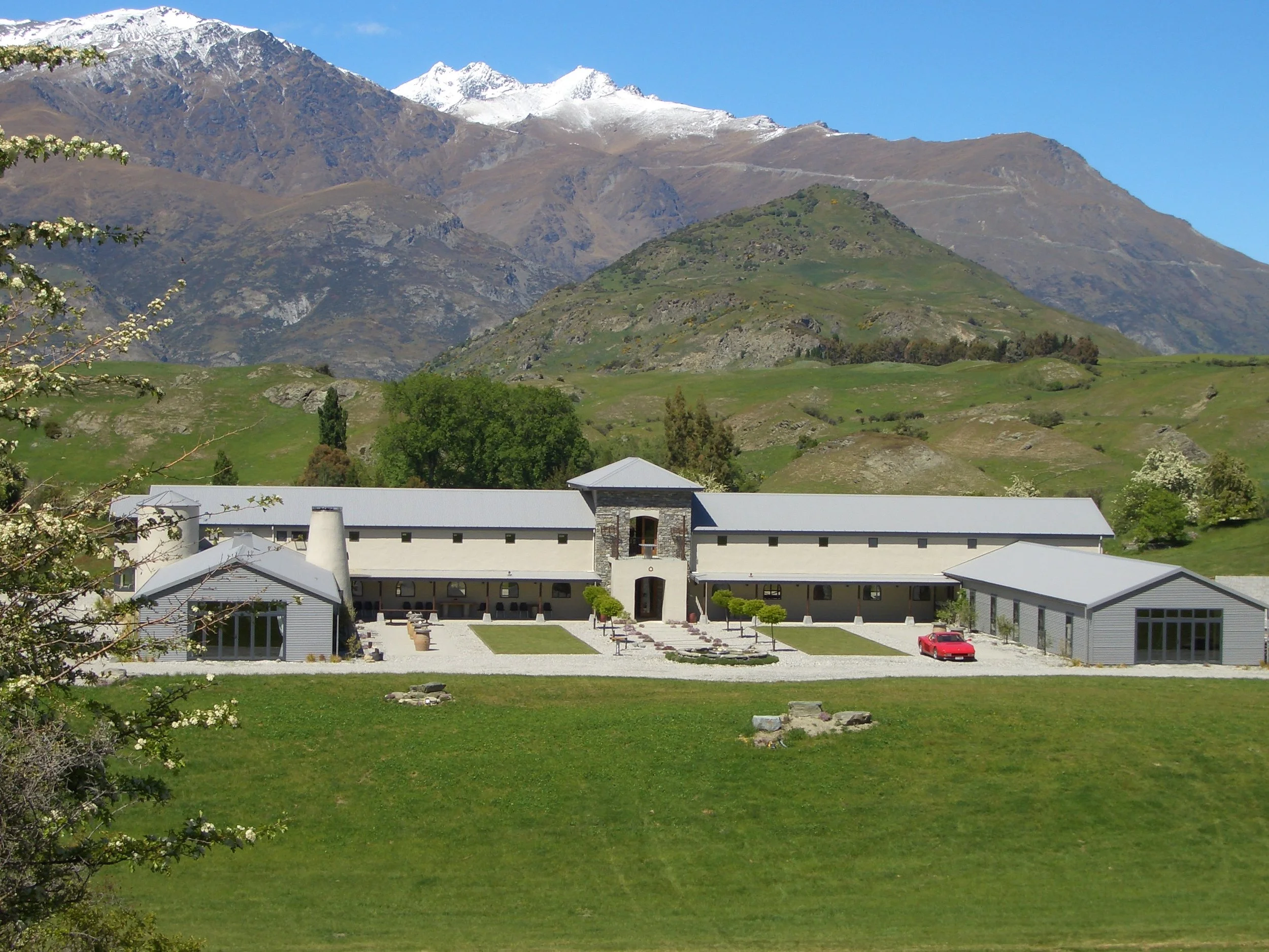 A large modern building complex with a central entrance, surrounded by green lawns and trees, with mountains in the background, some of which are snow-capped.