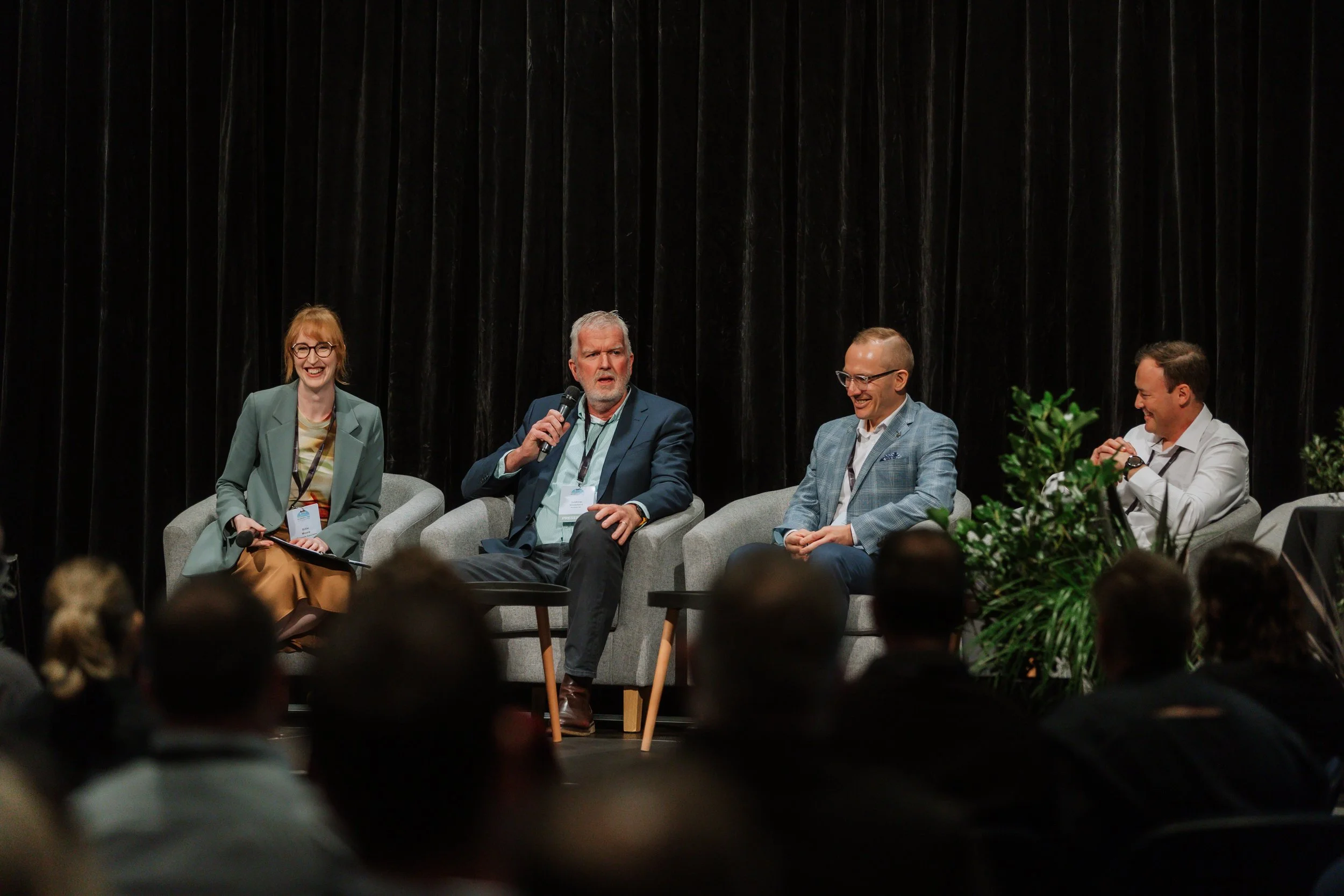 A panel discussion featuring four people seated on stage with dark curtains in the background, and an audience in front.