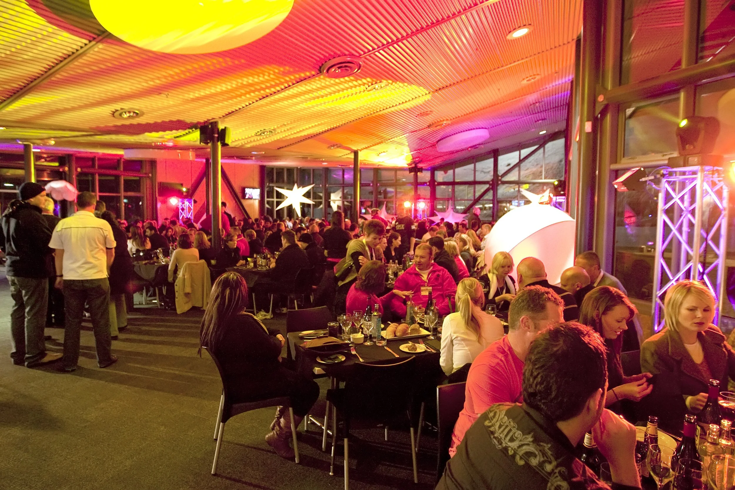 A crowded indoor event with many people seated at tables, celebrating with food and drinks, under colorful lighting and festive decorations.