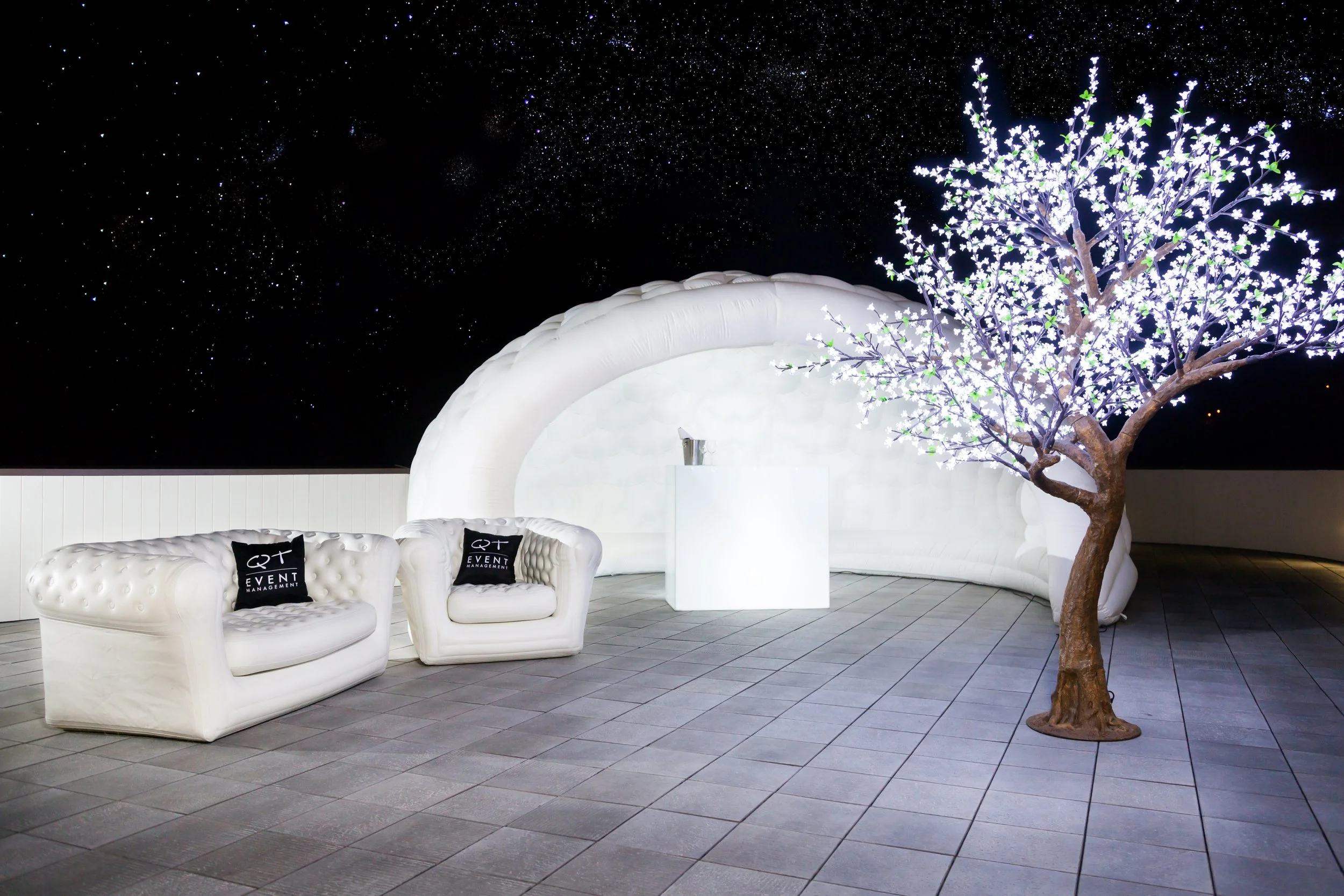 night view of a decorated outdoor event space with white cushioned chairs, a curved inflatable structure, a white square table with a bottle, a lit-up artificial tree, and a starry sky in the background.