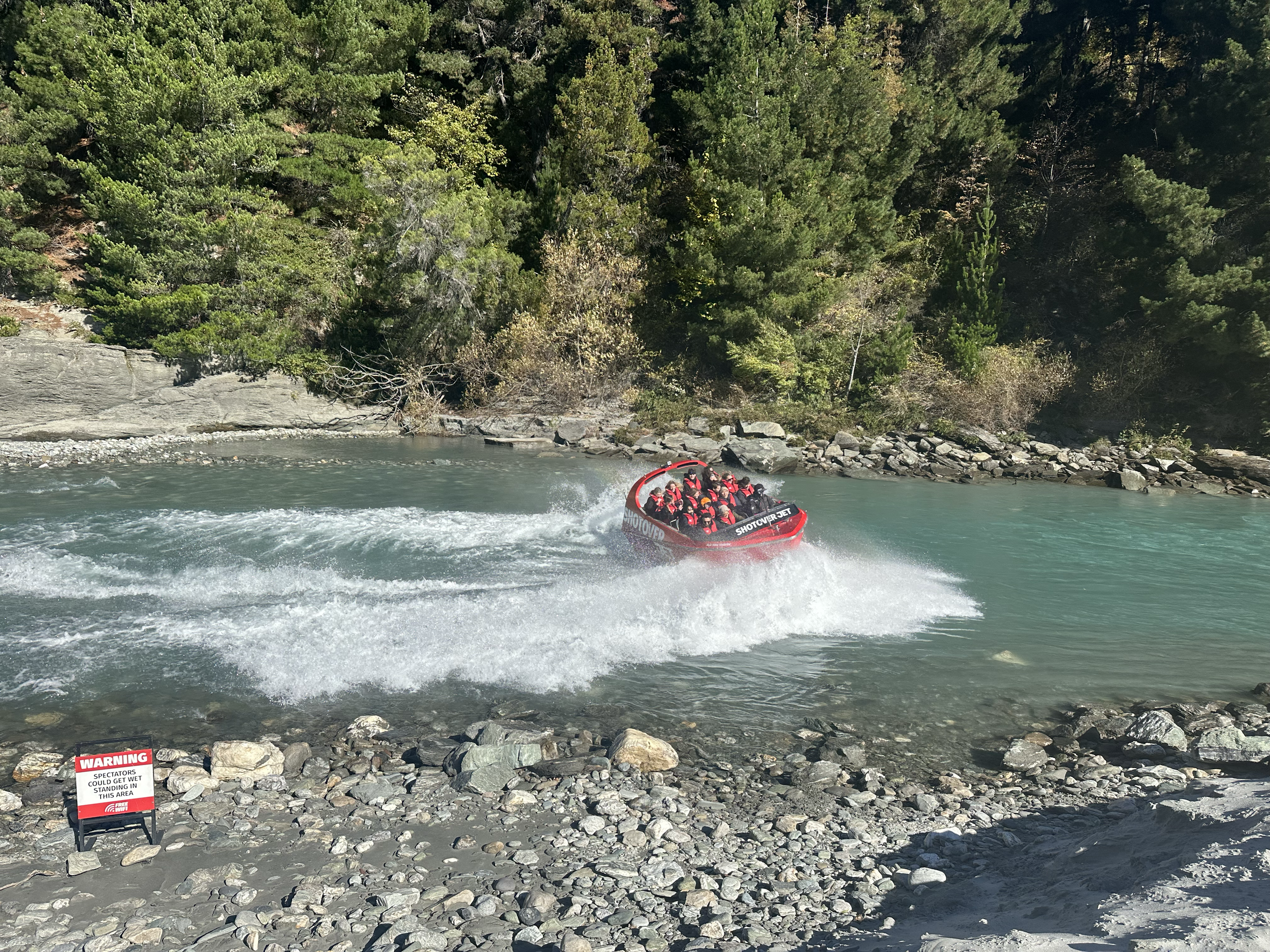 A group of people riding a red and black motorboat on a river surrounded by trees and rocks, with a warning sign on the shore indicating spectators could get wet.