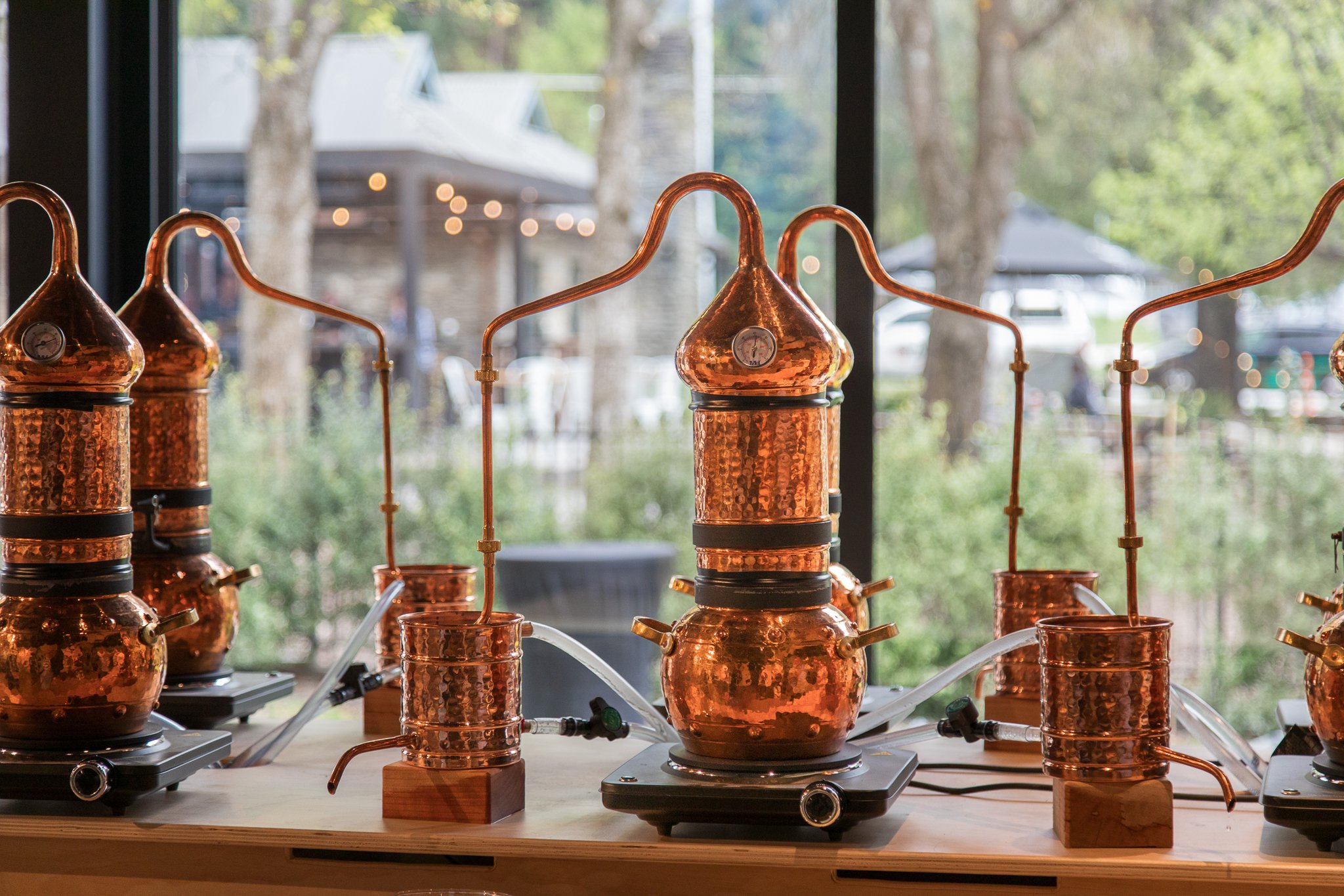 Copper distillation apparatus set up on a wooden surface, with a large copper pot connected to a glass condenser system, in front of a window with a blurred outdoor background.