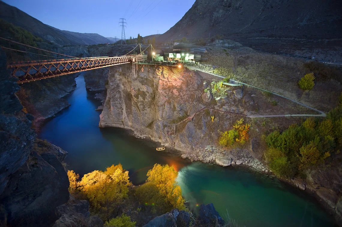 Night view of the Golden Gate Bridge in San Francisco, spanning a deep gorge with a river below, surrounded by hills with autumn foliage and faint city lights in the distance.