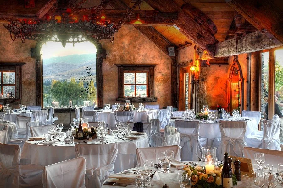 A rustic banquet hall decorated for a wedding reception with white-covered tables, glassware, and floral centerpieces, overlooking a mountain landscape through large windows.