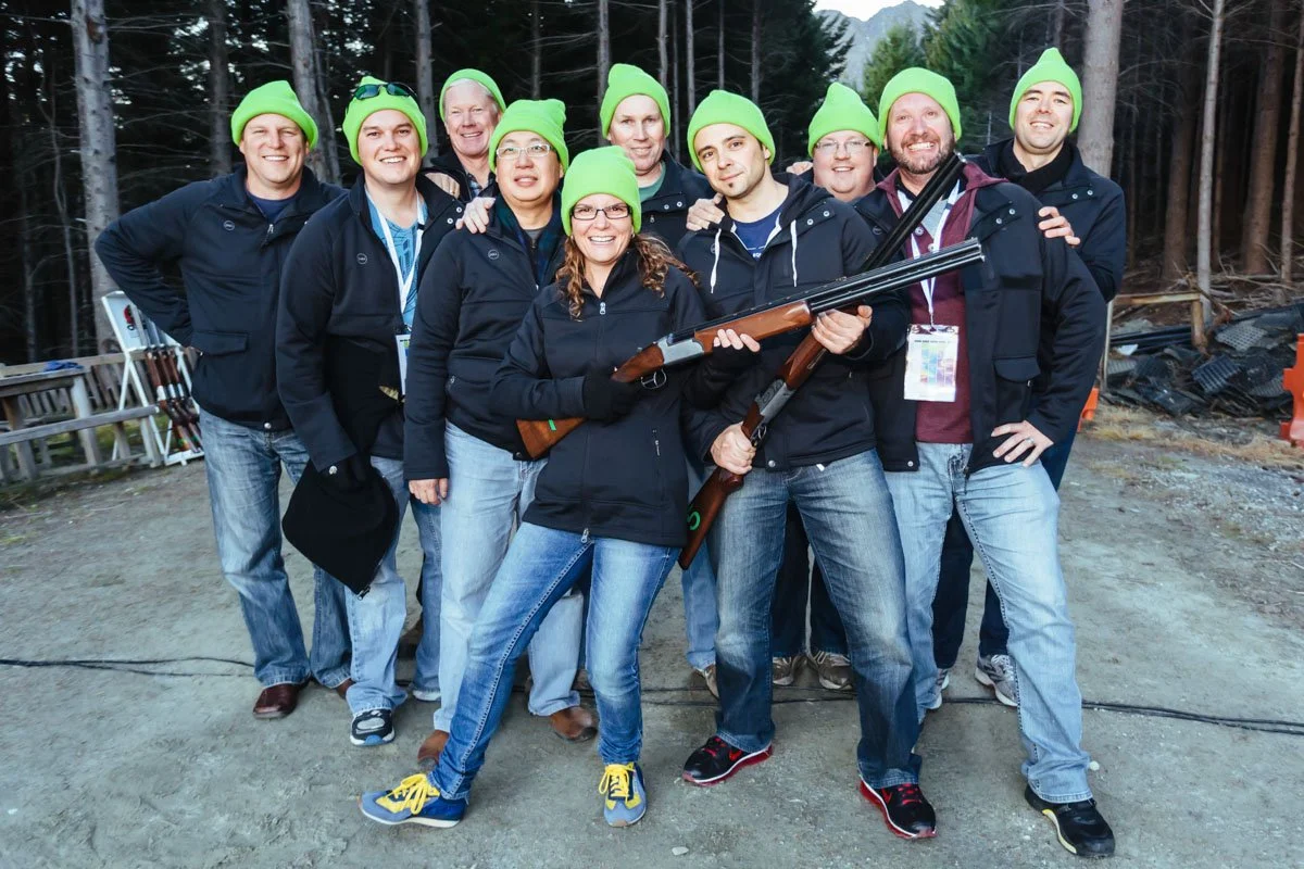Group of people outdoors wearing matching green beanies and black jackets, with two individuals in front holding shotguns, in a forested area.