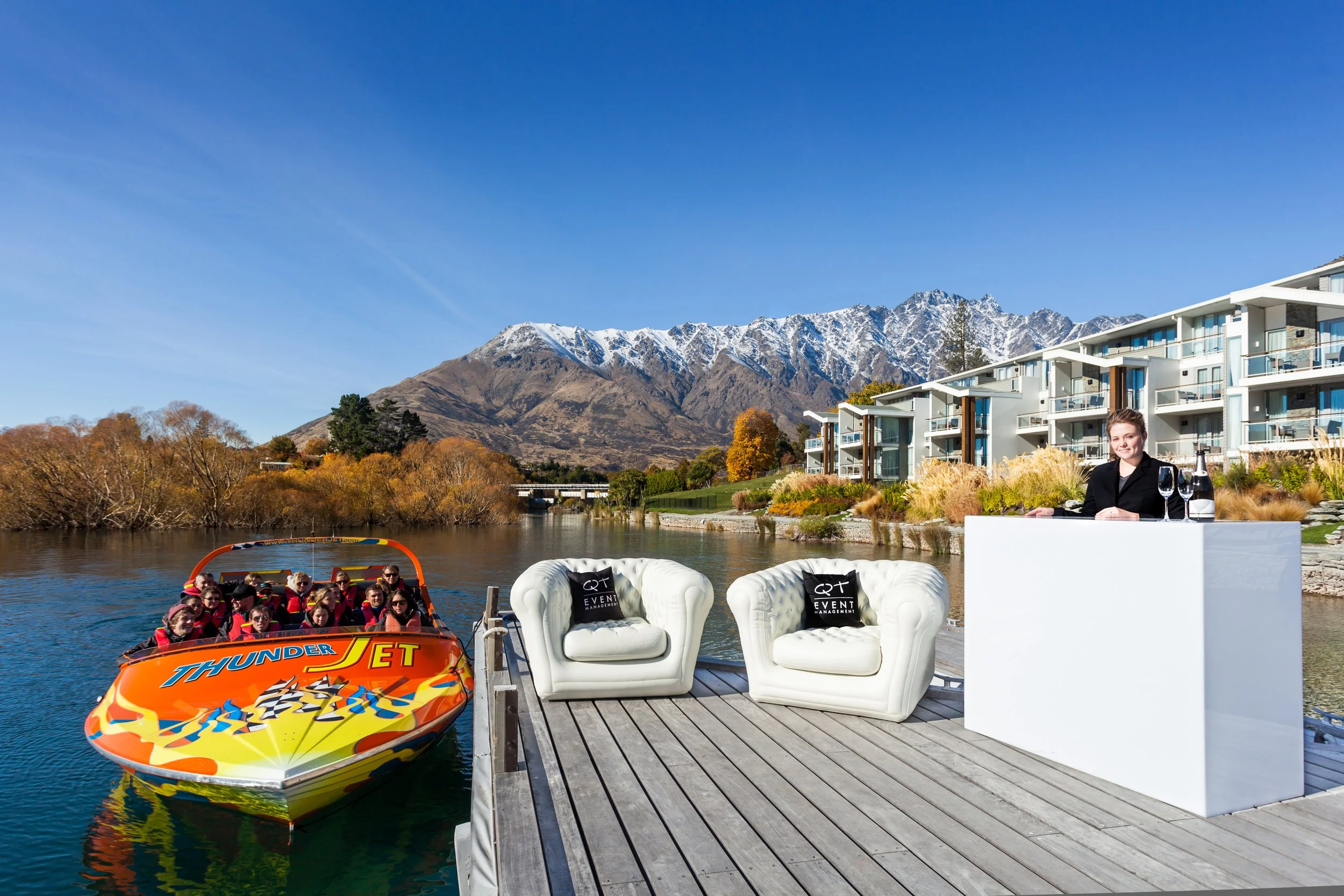 A dock on a river with two white chairs with black pillows, a woman standing behind a white counter with drinks, a speedboat on the water, modern apartment buildings, autumn trees, and snow-capped mountains under a clear blue sky.