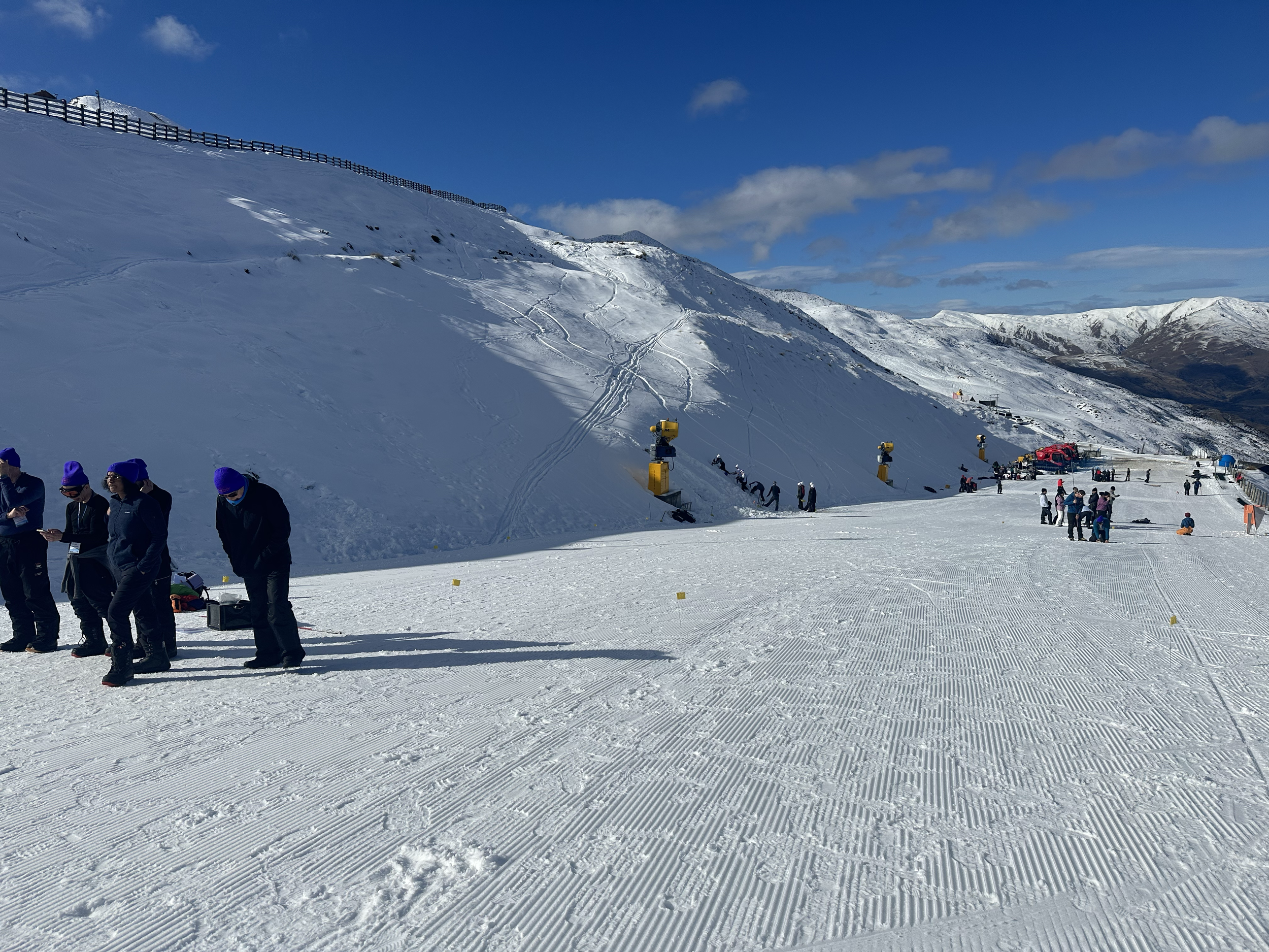 People dressed in winter clothing and blue hats stand on snow at a ski resort with ski slopes and snow-covered mountains in the background.