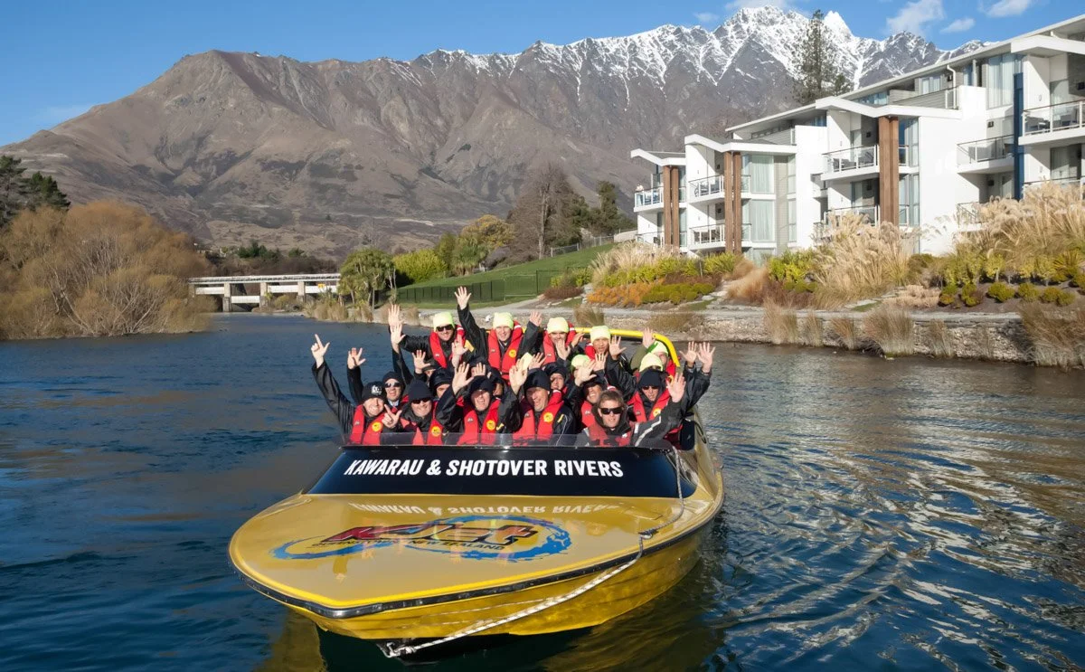 Group of people in a yellow boat wearing safety jackets and helmets, waving and smiling, on a river with mountains in the background.