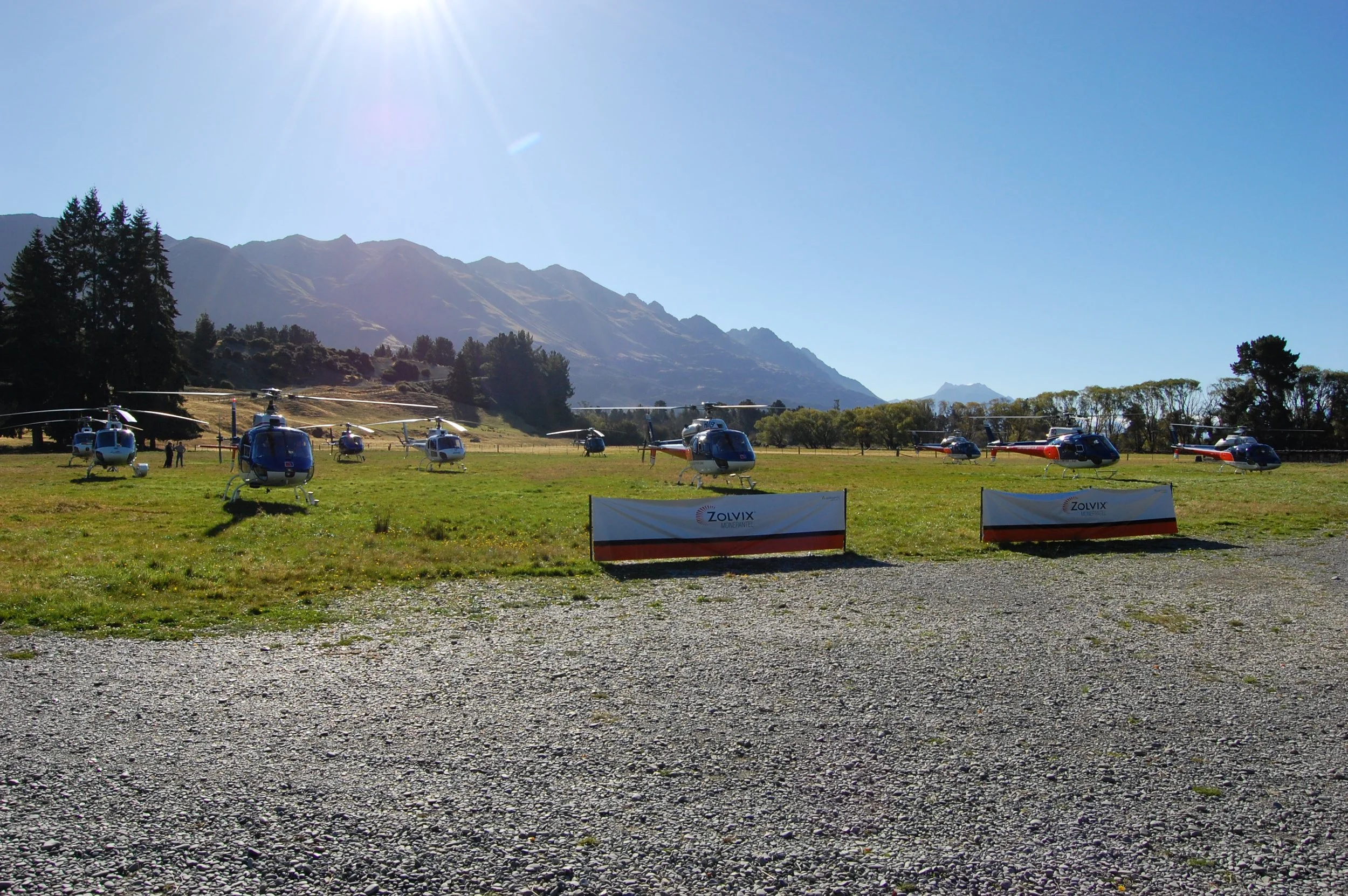 Multiple helicopters parked on a grassy field with mountains in the background and a clear blue sky.