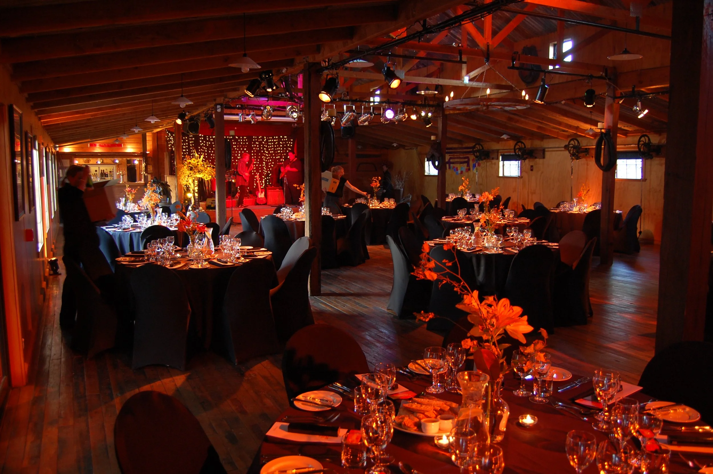 Elegant banquet hall with round tables covered in black tablecloths, decorated with floral centerpieces, set with glassware, plates, and silverware, featuring a wooden floor and a stage with live musicians under warm lighting.