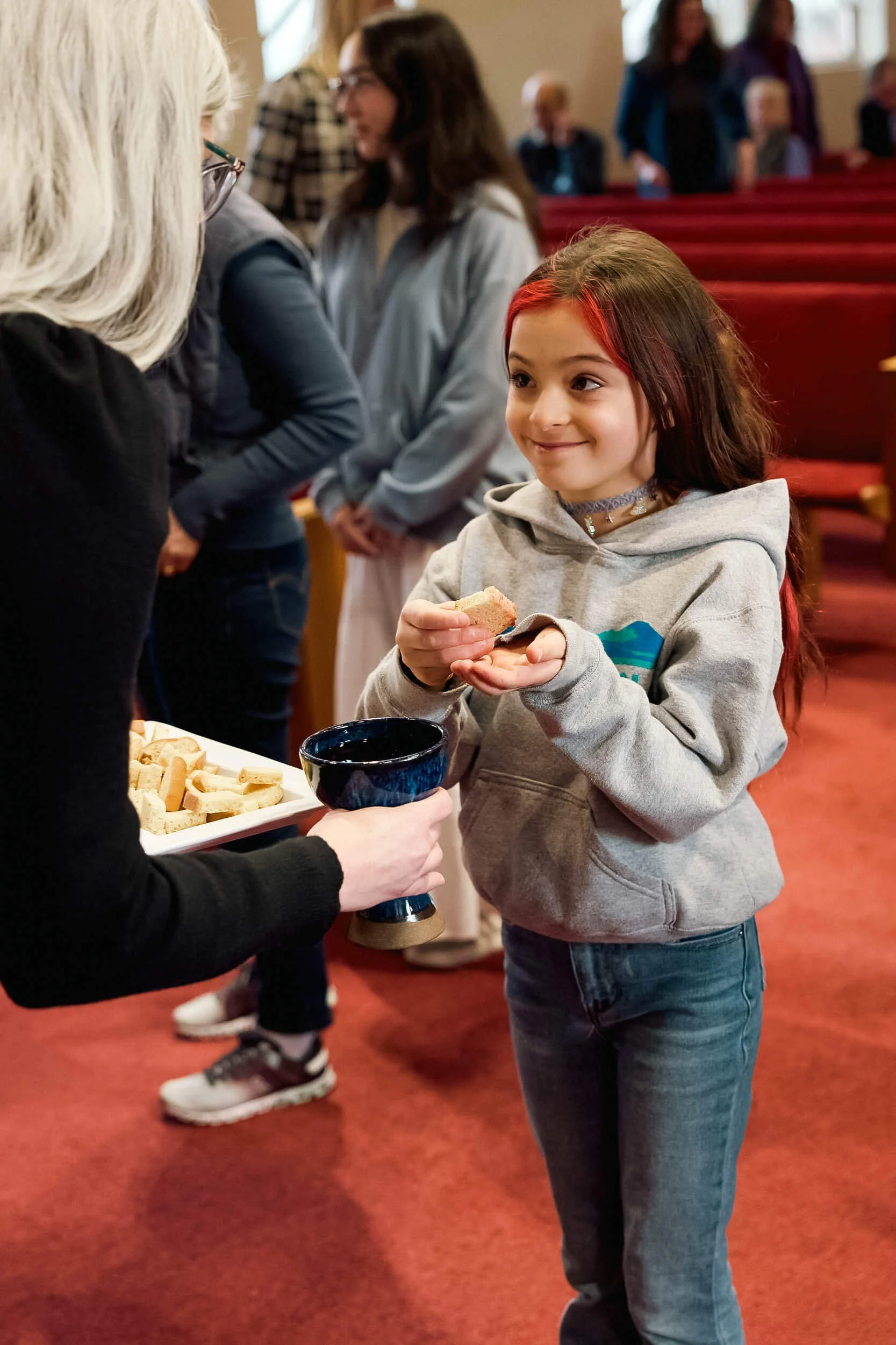 A young girl with red highlights receives communion with a smile as the pastor leans down to meet her at her level during a worship service at Crown Hill Church.