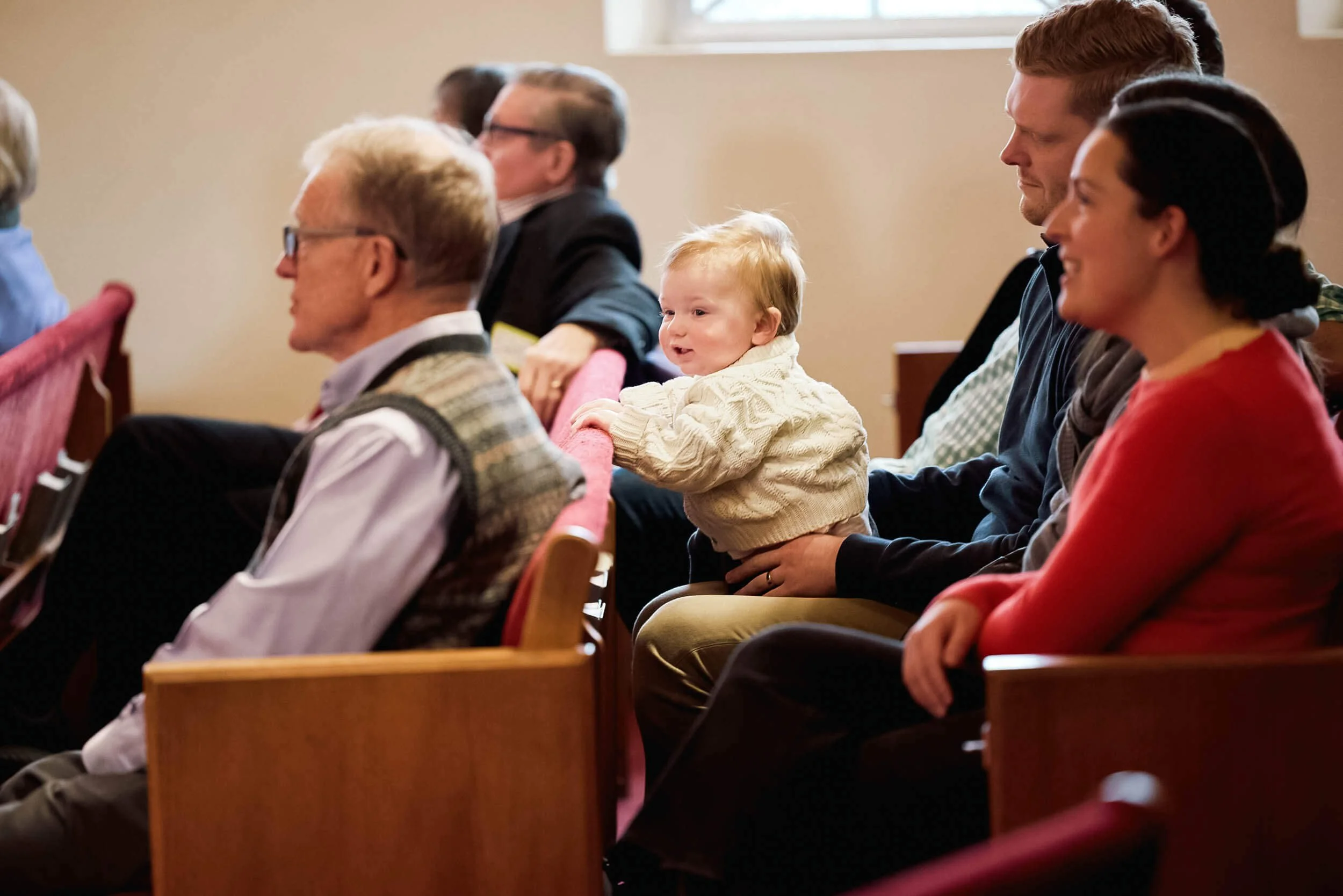A baby sits on a parent's lap gripping the pew ahead, while an older gentleman sits in the row in front, capturing the multigenerational community at Crown Hill Church in Seattle's Crown Hill neighborhood.