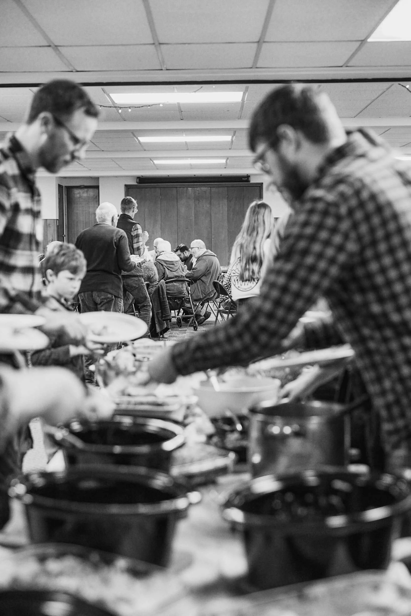 A black and white image of a church potluck at Crown Hill Church, with blurred figures serving themselves food in the foreground and tables of people eating and conversing together in the background in Seattle's Crown Hill neighborhood.