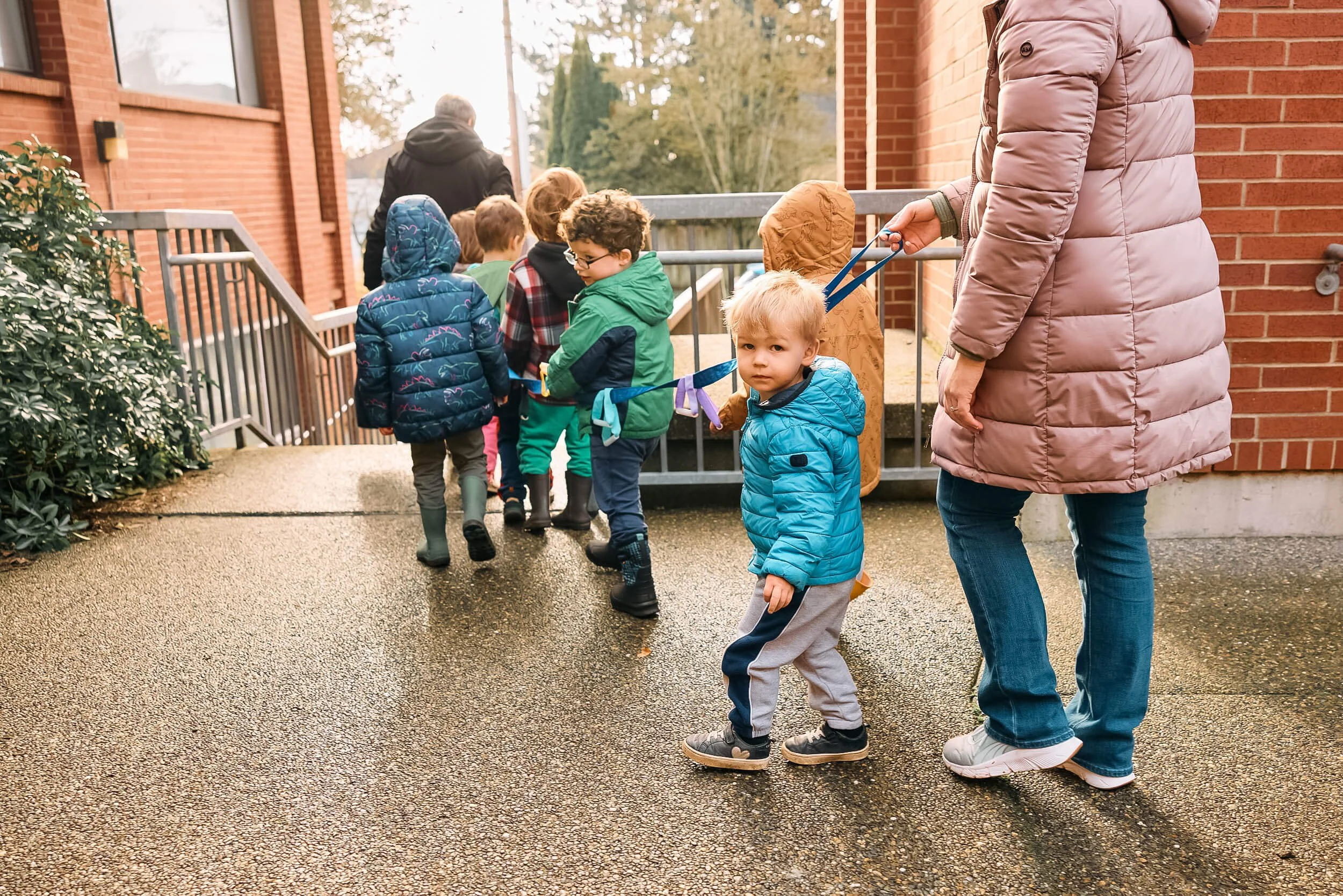 A group of preschool children walk together holding a blue fabric chain while a teacher follows close behind, with one child looking directly at the camera, at PATH Preschool at Crown Hill Church in Seattle's Crown Hill neighborhood.