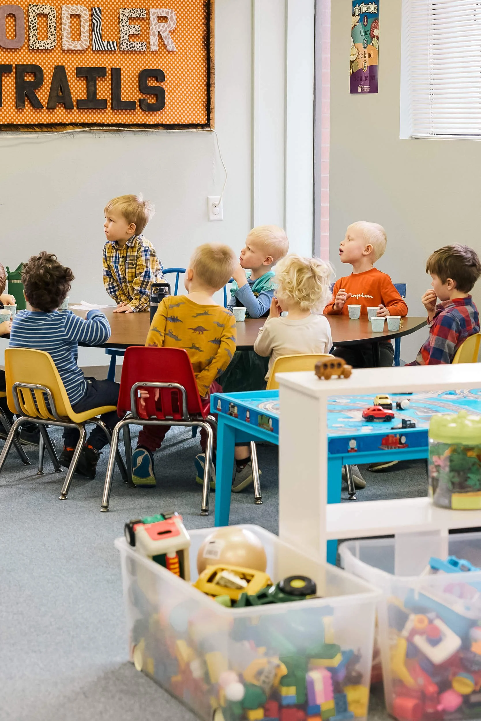 Preschool aged children sit at a table eating snack and listening to their teacher during Sunday school.