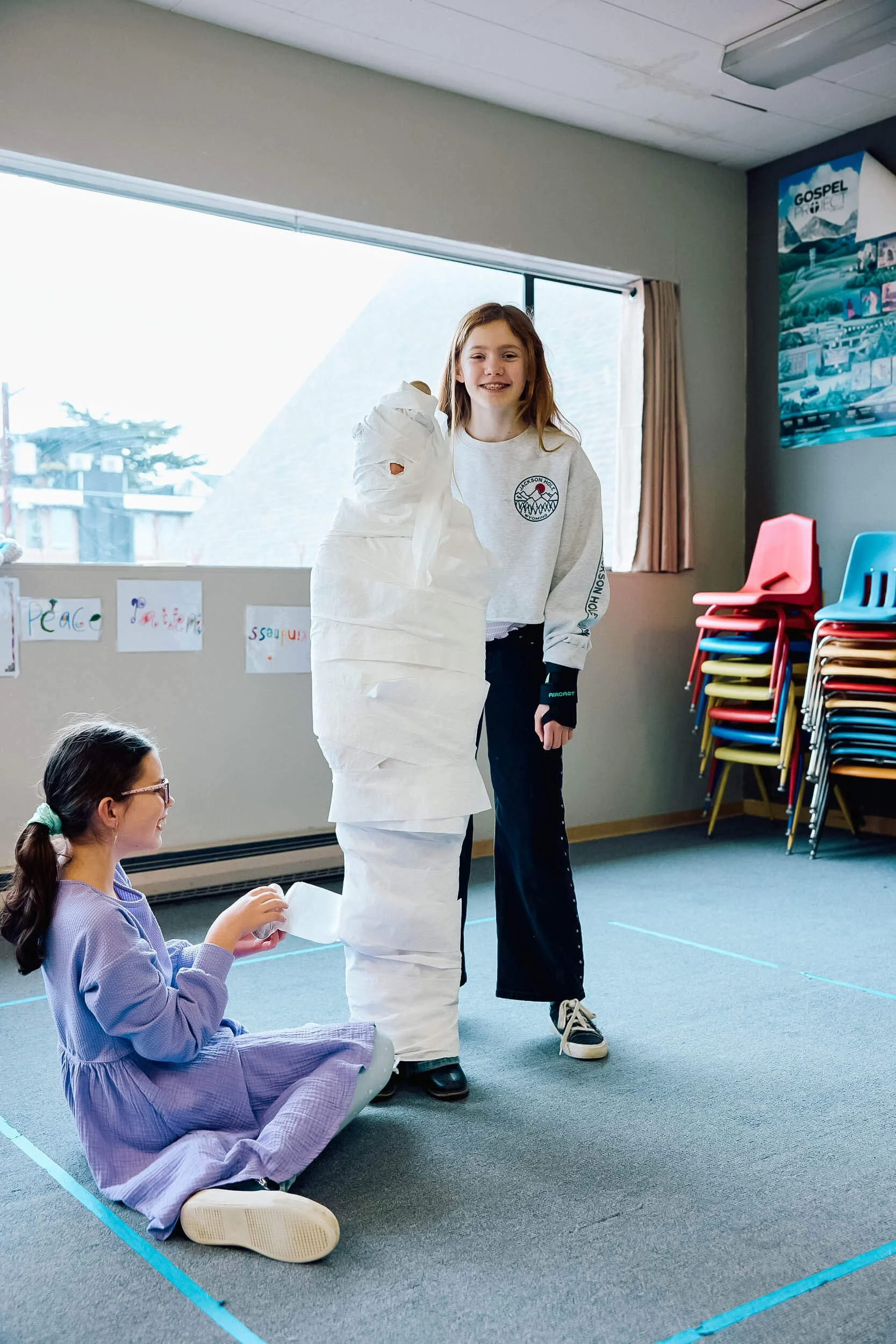 Two tween girls laugh and work together wrapping a child in toilet paper during a fun game at Sunday school at Crown Hill Church in Seattle's Crown Hill neighborhood.