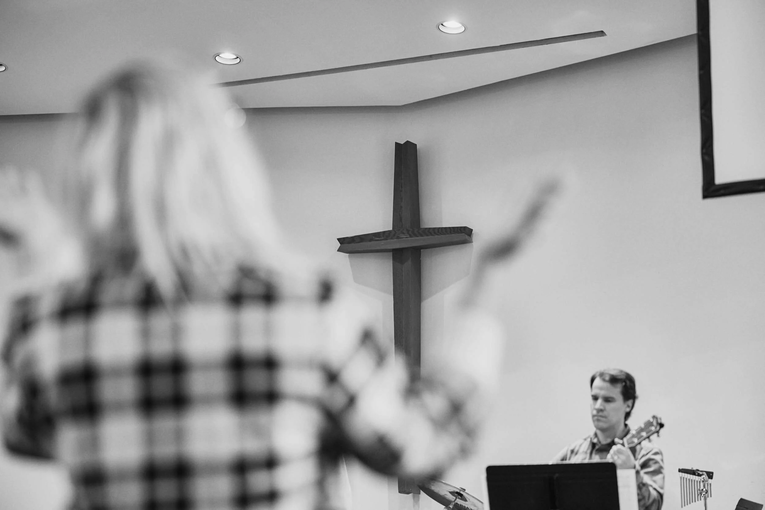 The sanctuary cross in focus at Crown Hill Church as a woman raises her hands in worship and a guitarist leads from the stage, welcoming all who have questions about faith and community.