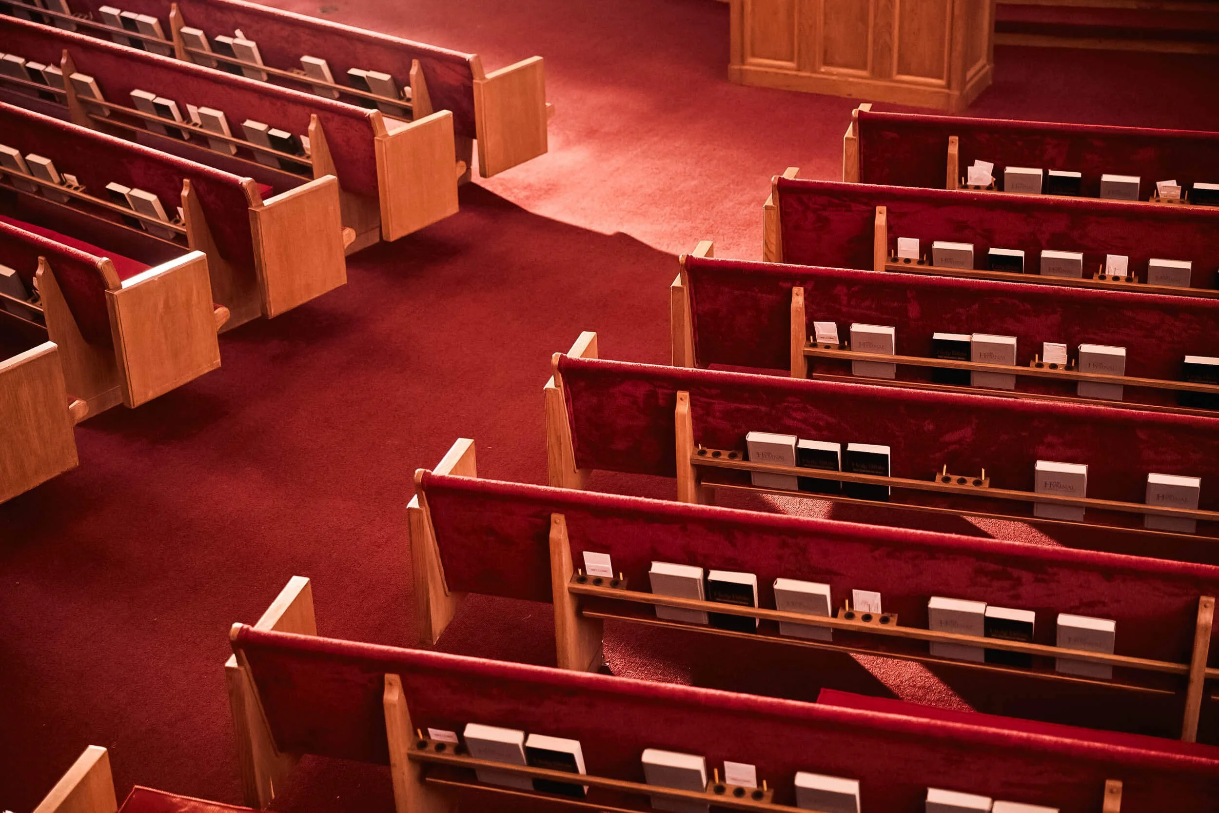 Empty church pews with red velvet upholstery and wooden armrests, arranged in rows on a red carpeted floor
