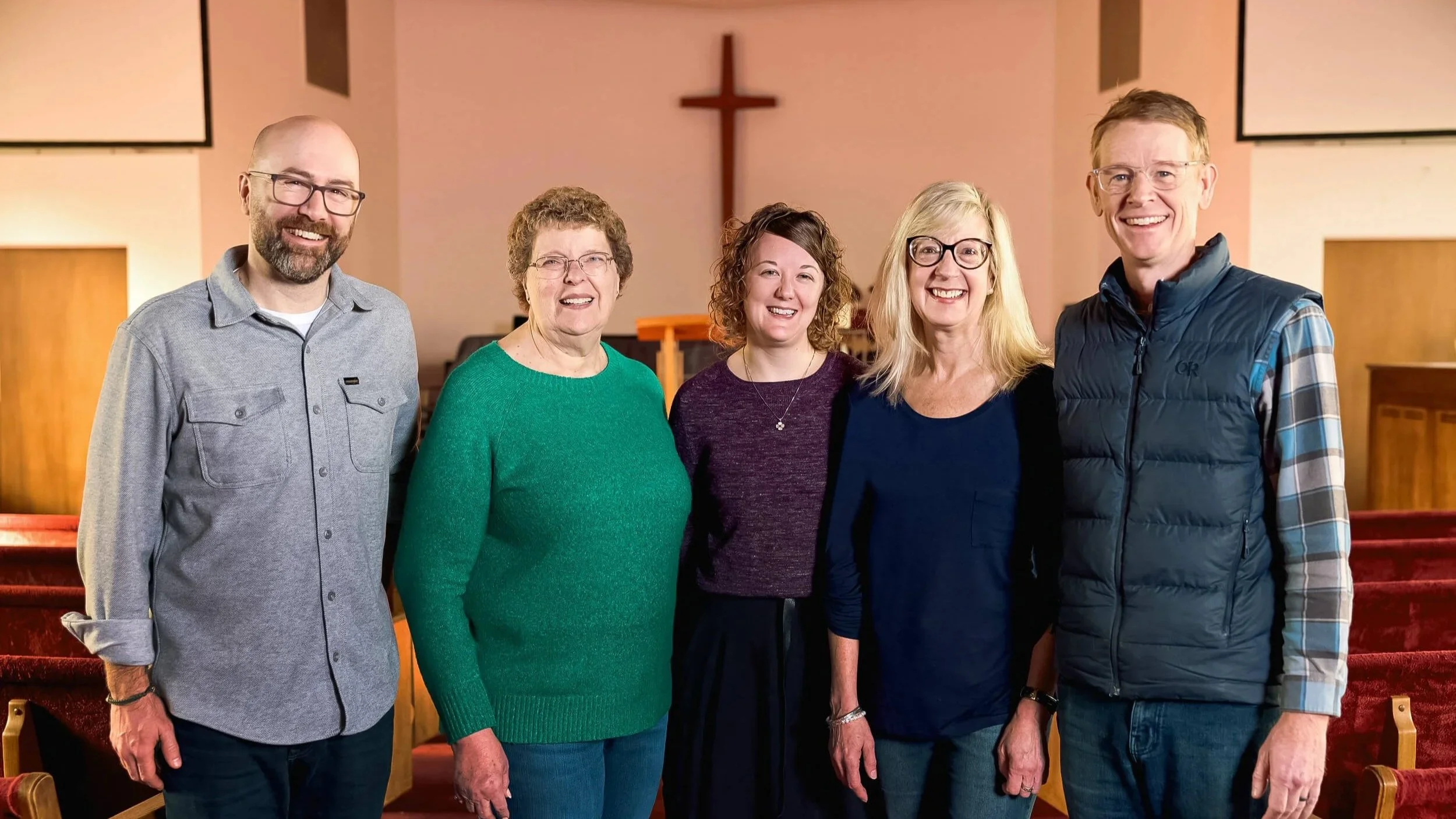 Five Crown Hill Church staff members stand shoulder to shoulder, smiling warmly at the camera with the sanctuary cross softly blurred behind them.