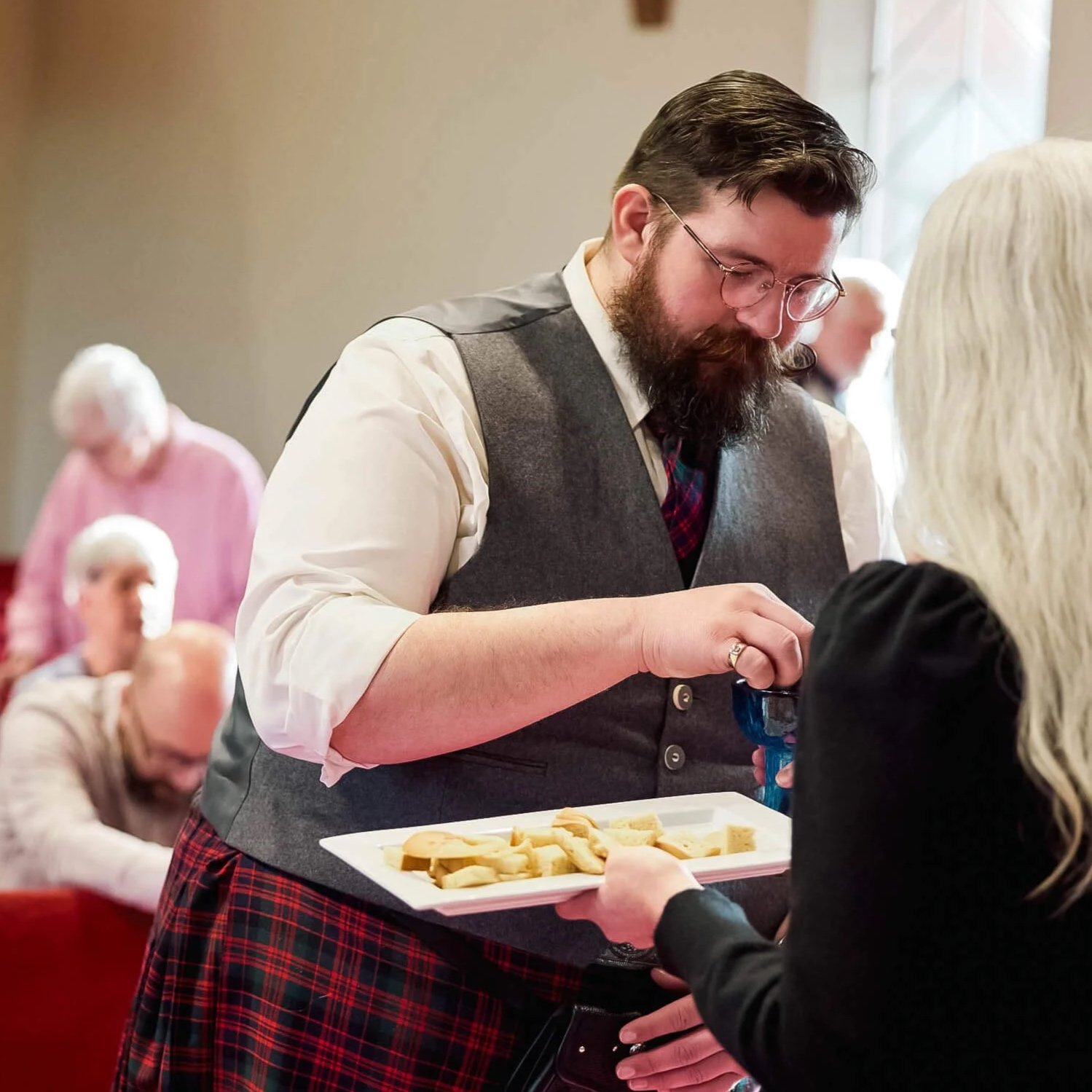 A man in a Scottish kilt receives communion while congregation members pray and worship in the blurred background at Crown Hill Church.