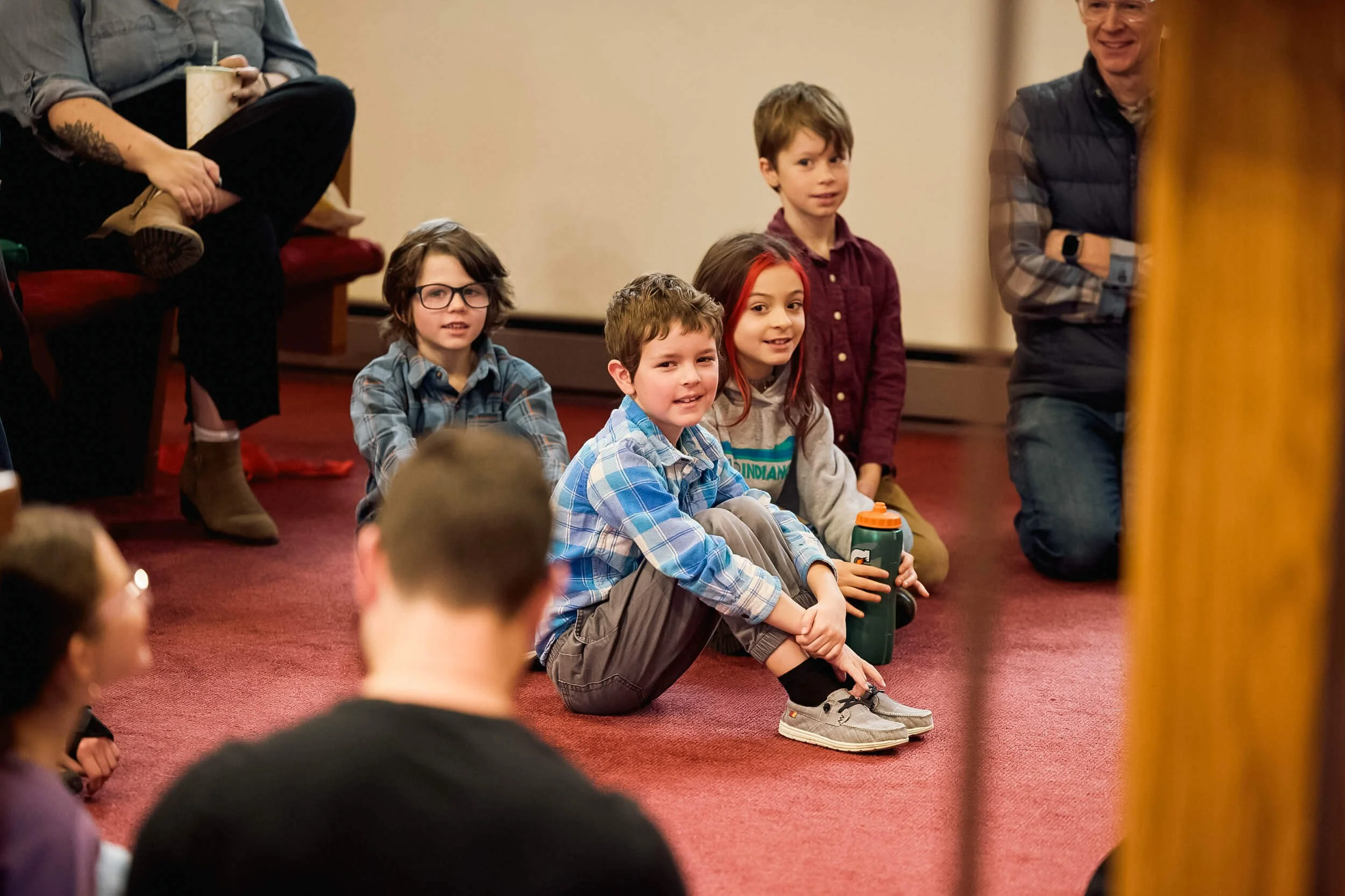 Four children sit on the floor at the front of the sanctuary with smiles on their faces, engaged with the speaker during the Kids Moment at Crown Hill Church.