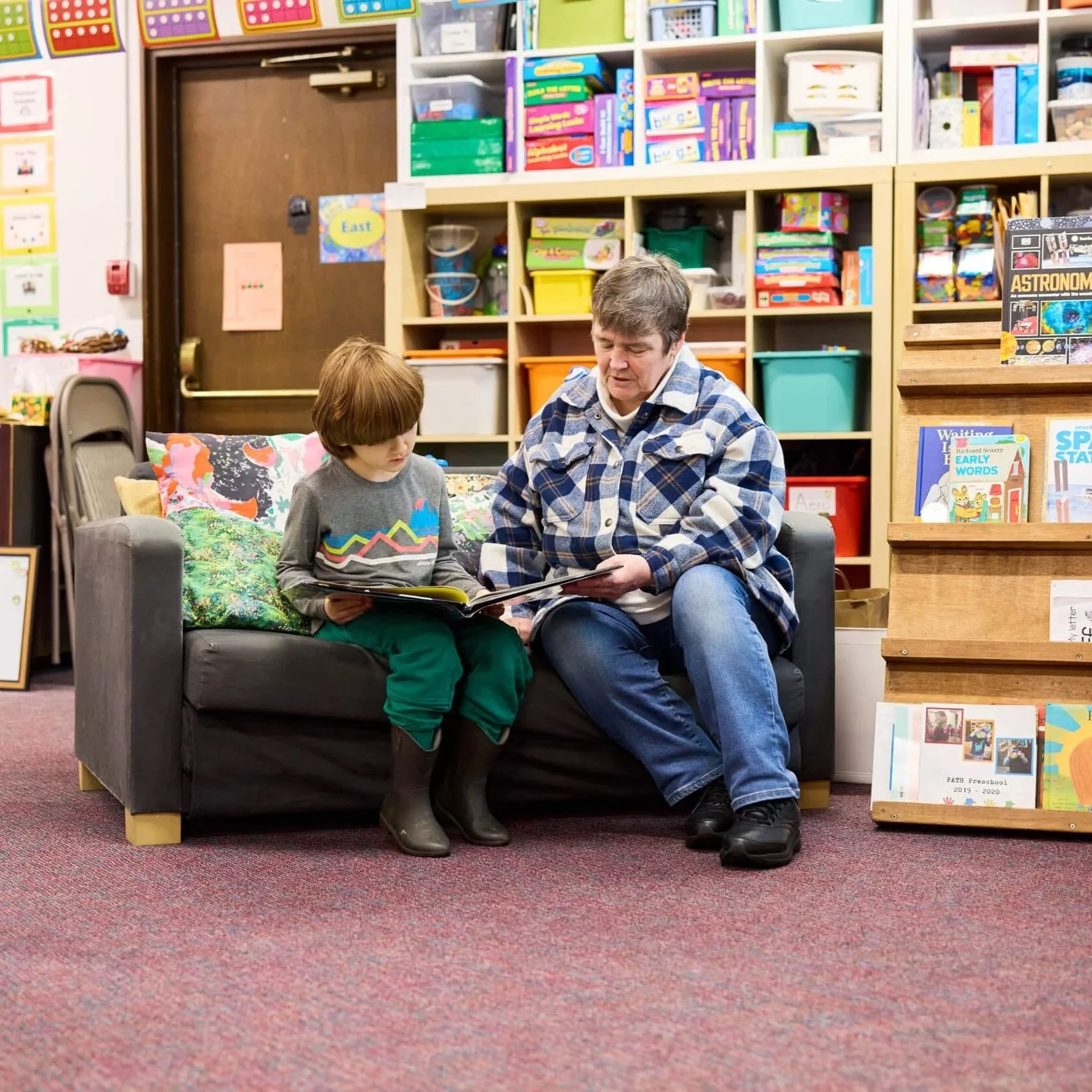 A teacher and young boy sit side by side on a couch, reading a book together at PATH Preschool.
