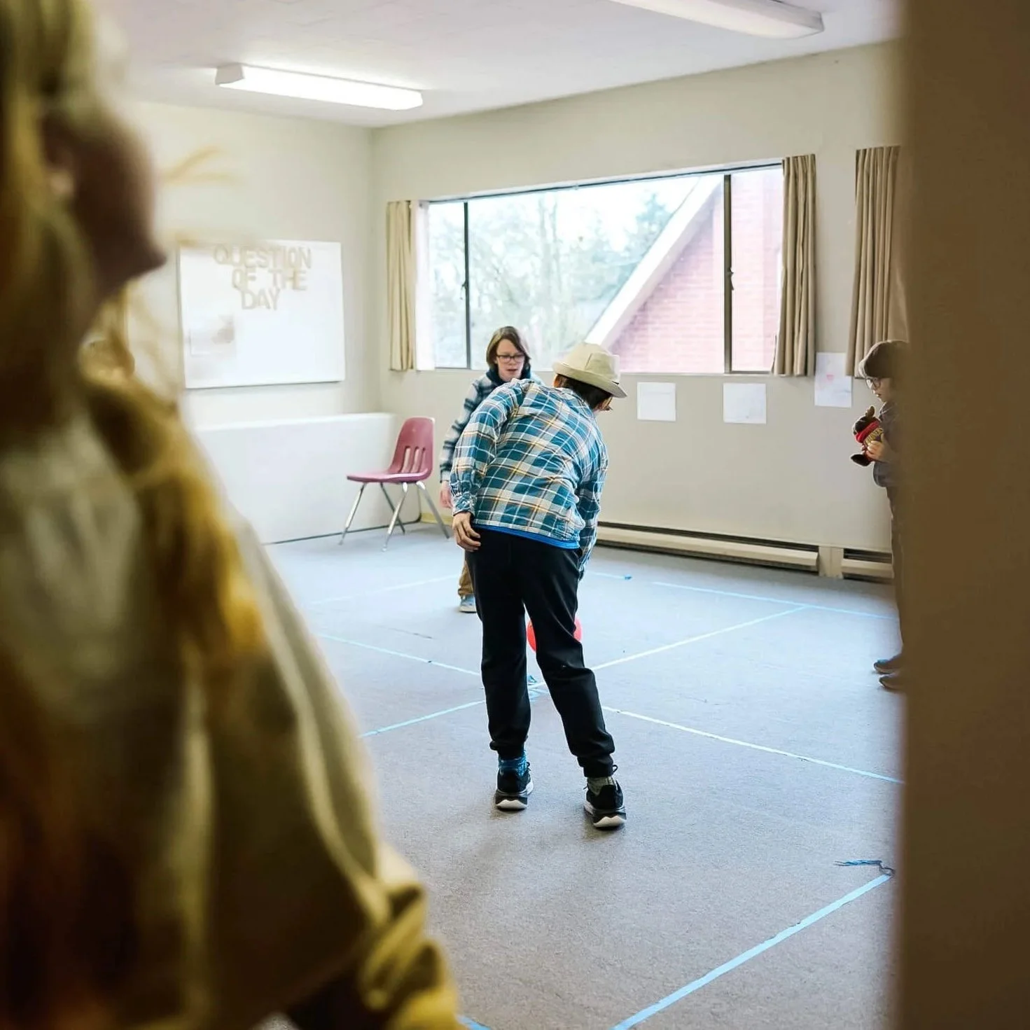 Three boys play four-square together viewed through an open doorway, while a girl watches from the foreground, during Sunday school at Crown Hill Church.