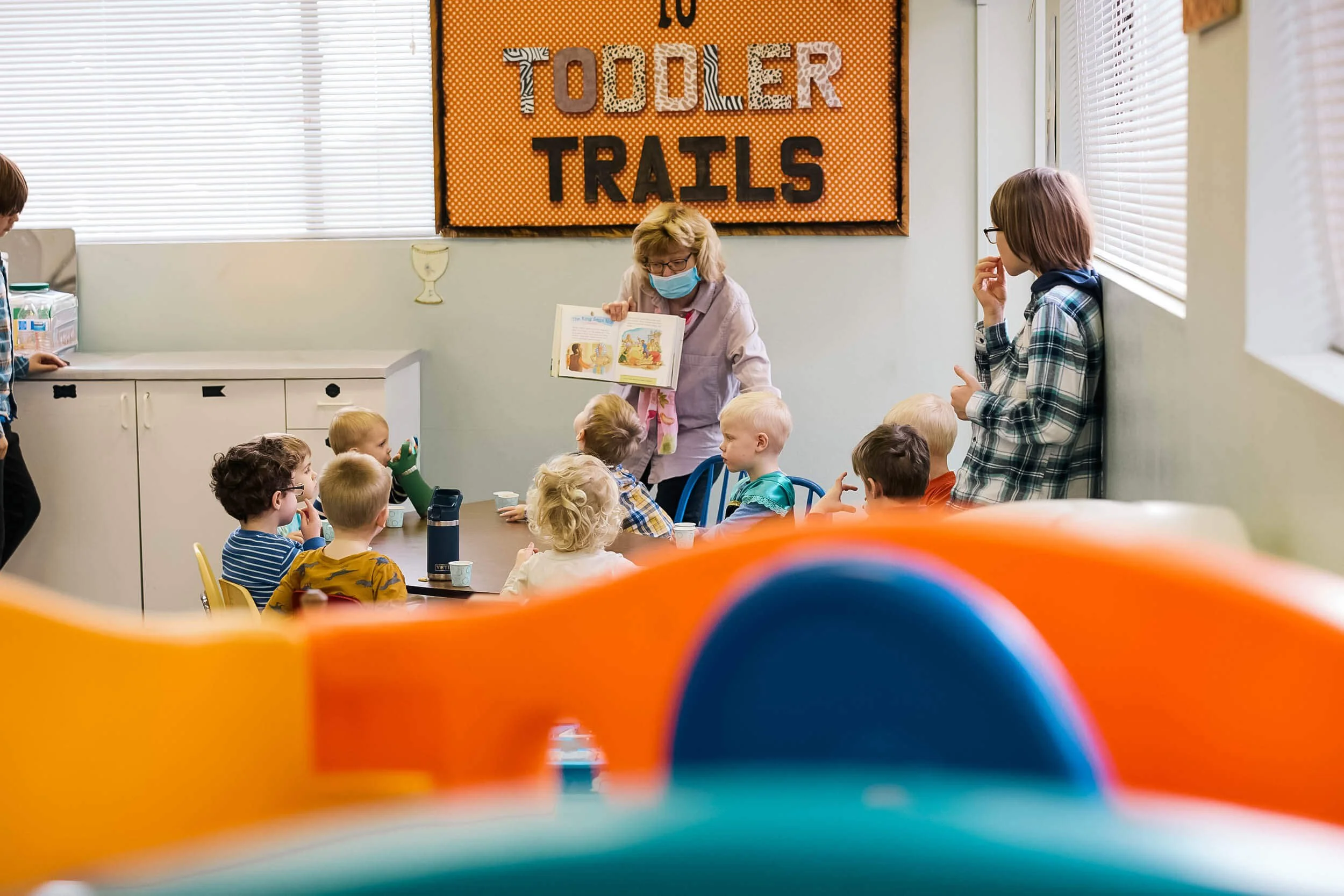 Preschoolers sit engaged and attentive as their masked teacher reads a Bible story in the Toddler Trails worship space at Crown Hill Church.