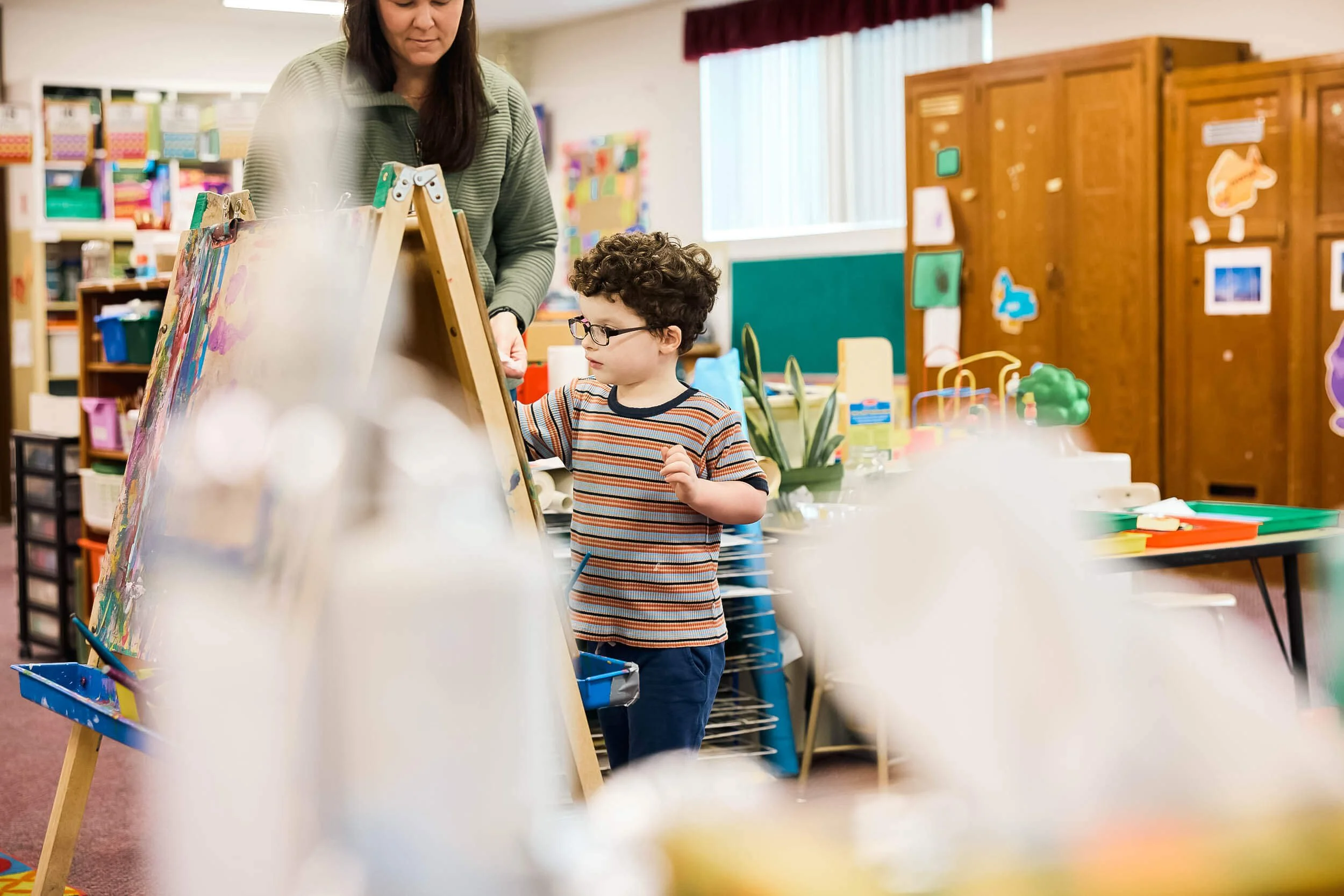 A boy paints at an easel with a teacher standing nearby at PATH Preschool at Crown Hill Church in Seattle's Crown Hill neighborhood.