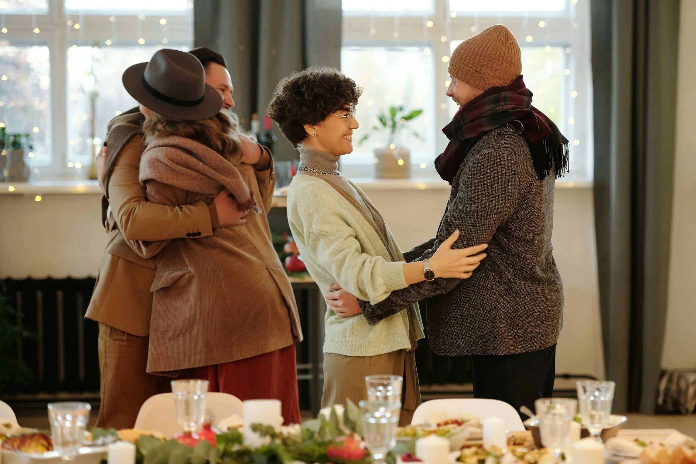 Four people gather warmly for a meal, two embracing in a hug and two greeting each other with smiles, representing the community meal groups that meet throughout Seattle connected to Crown Hill Church.