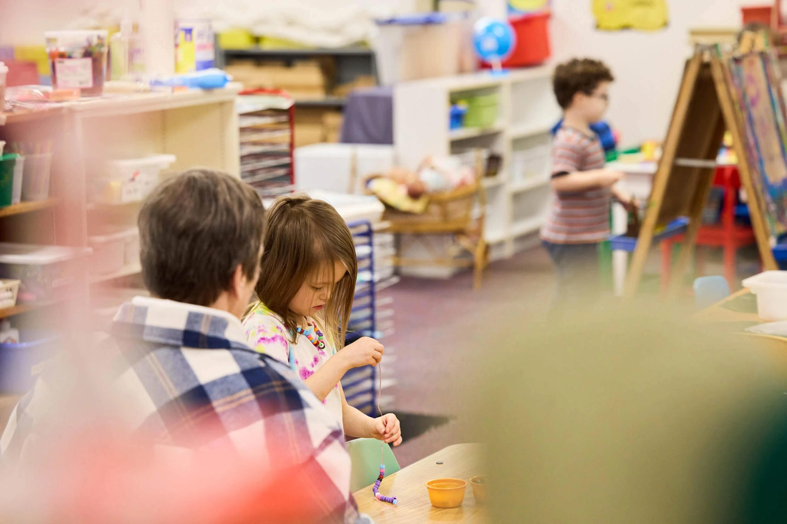 A teacher and a young girl work together on a beading activity at PATH Preschool, while a boy paints at an easel in the background.