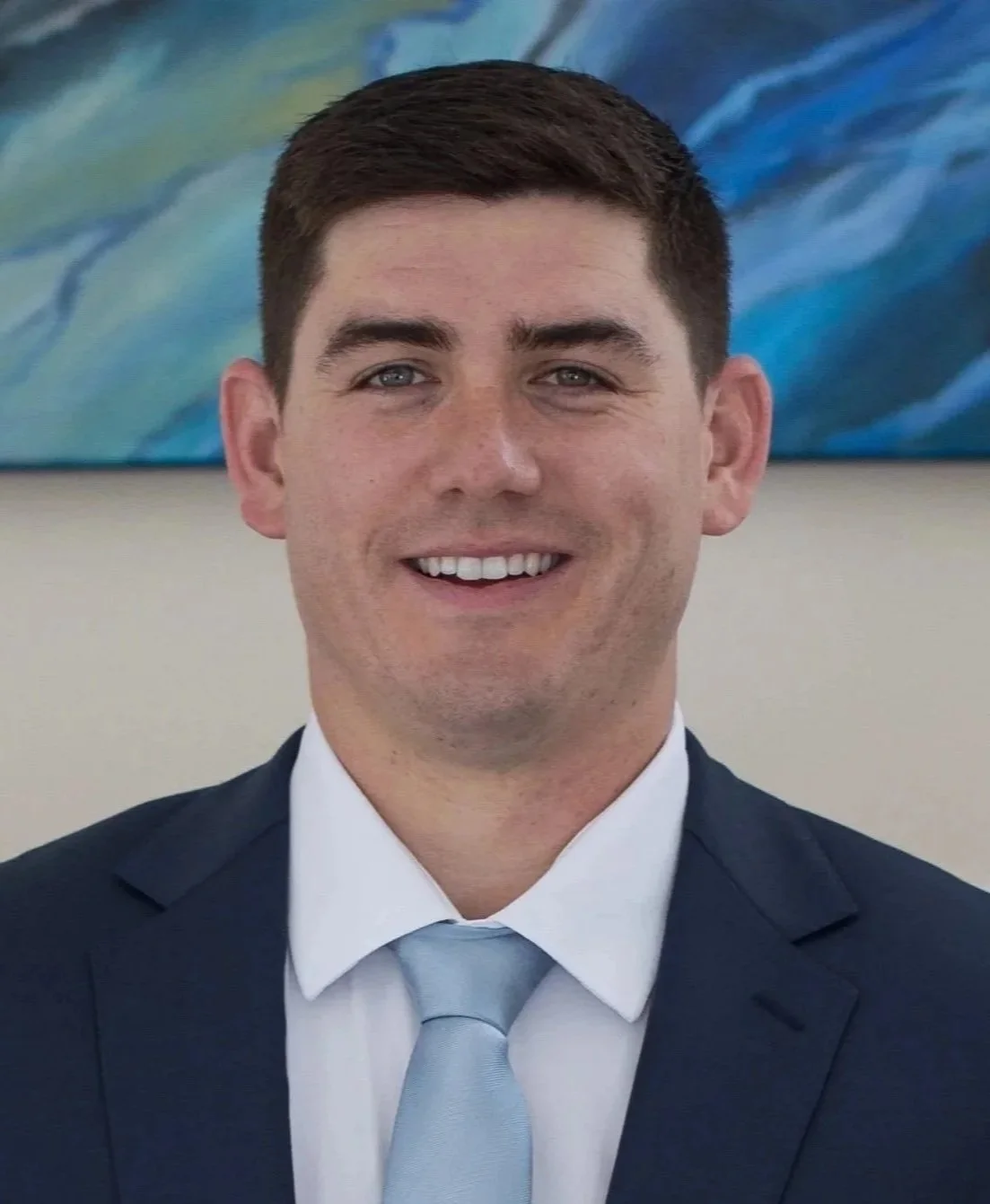 Young man in formal attire, smiling at the camera, with a blue abstract painting in the background.