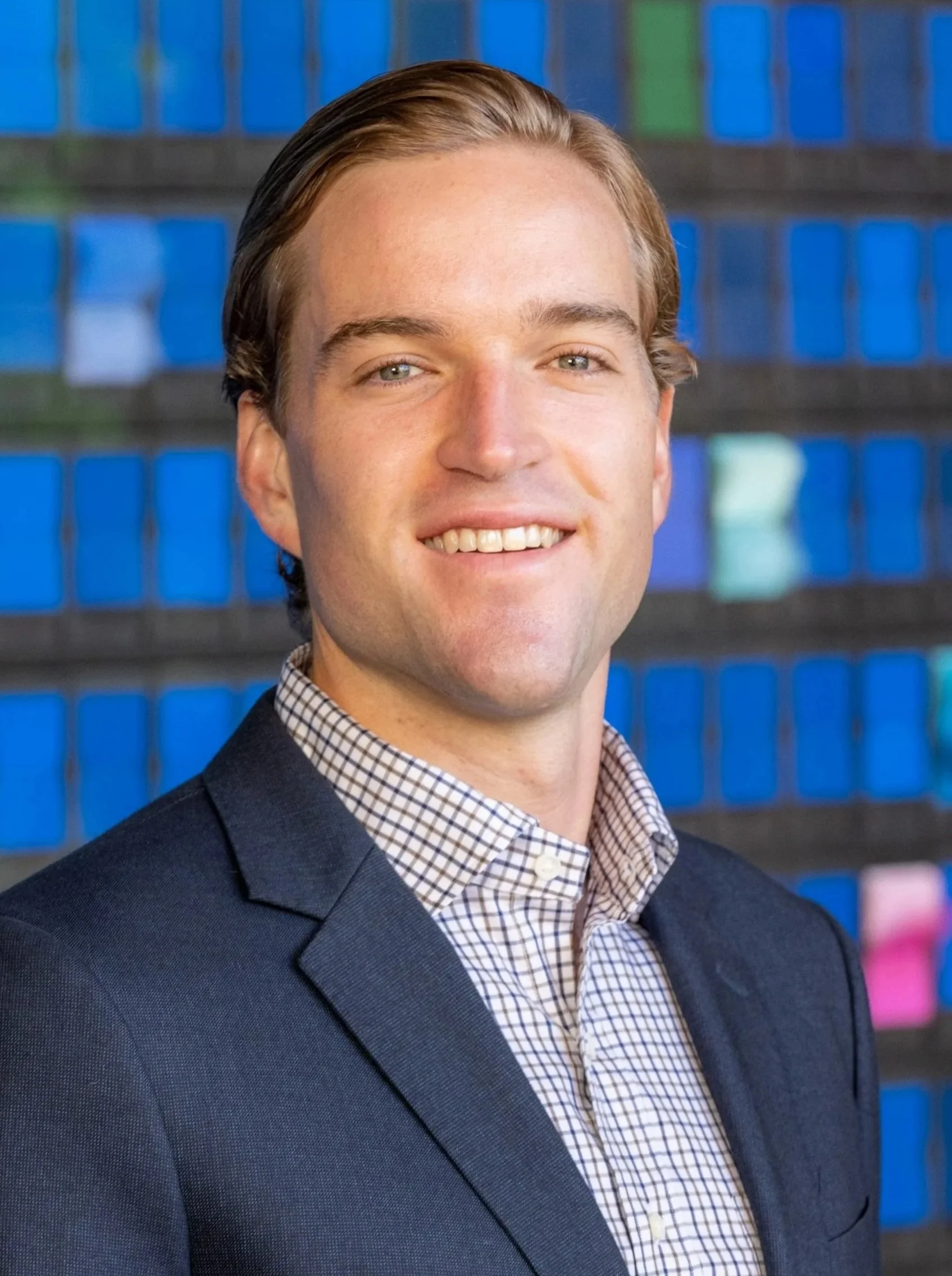 Headshot of a young man with light brown hair, wearing a dark suit jacket and a checkered dress shirt, smiling in front of a blue and green grid background.