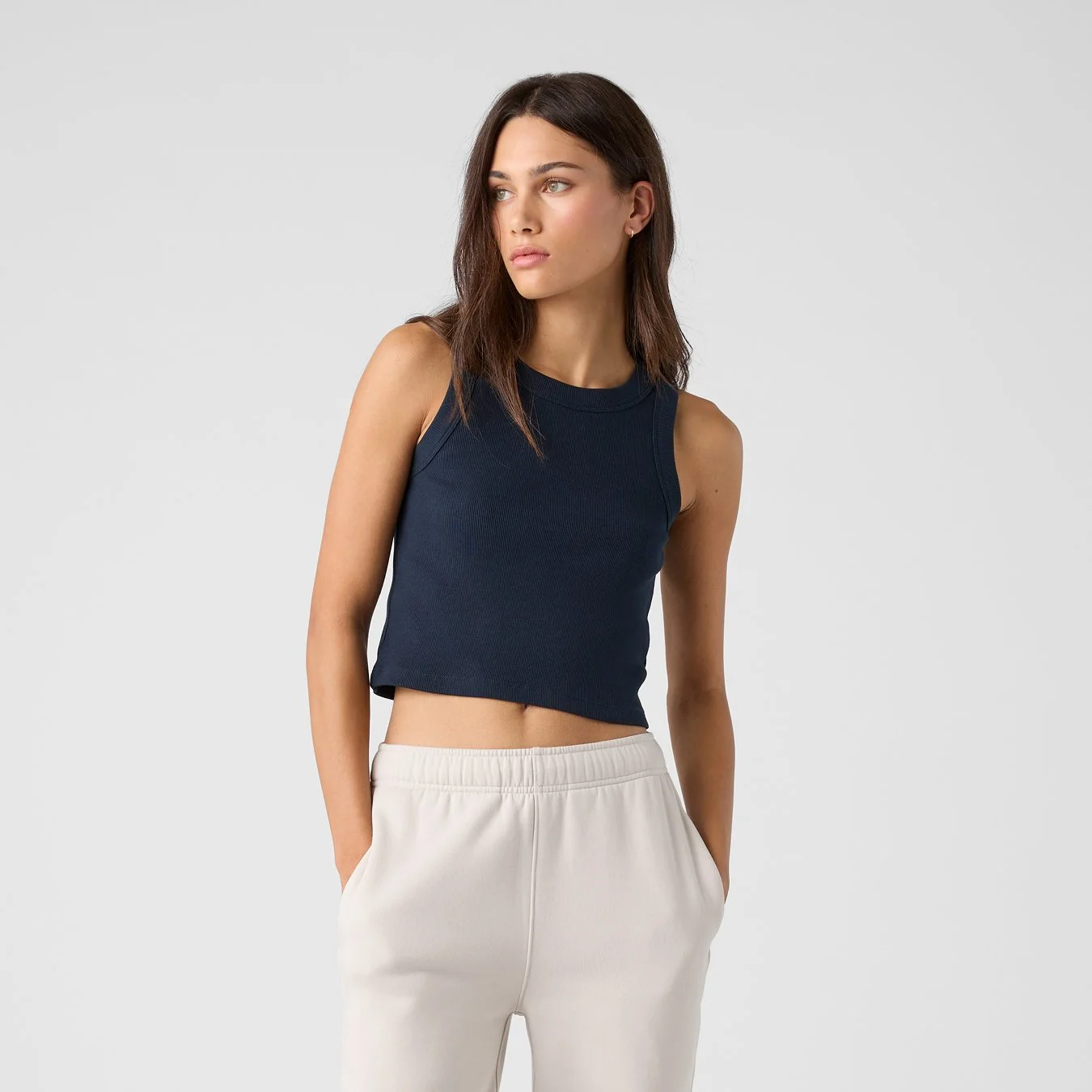 A young woman with shoulder-length dark hair and fair skin wearing a sleeveless navy crop top and beige sweatpants standing against a plain light gray background.