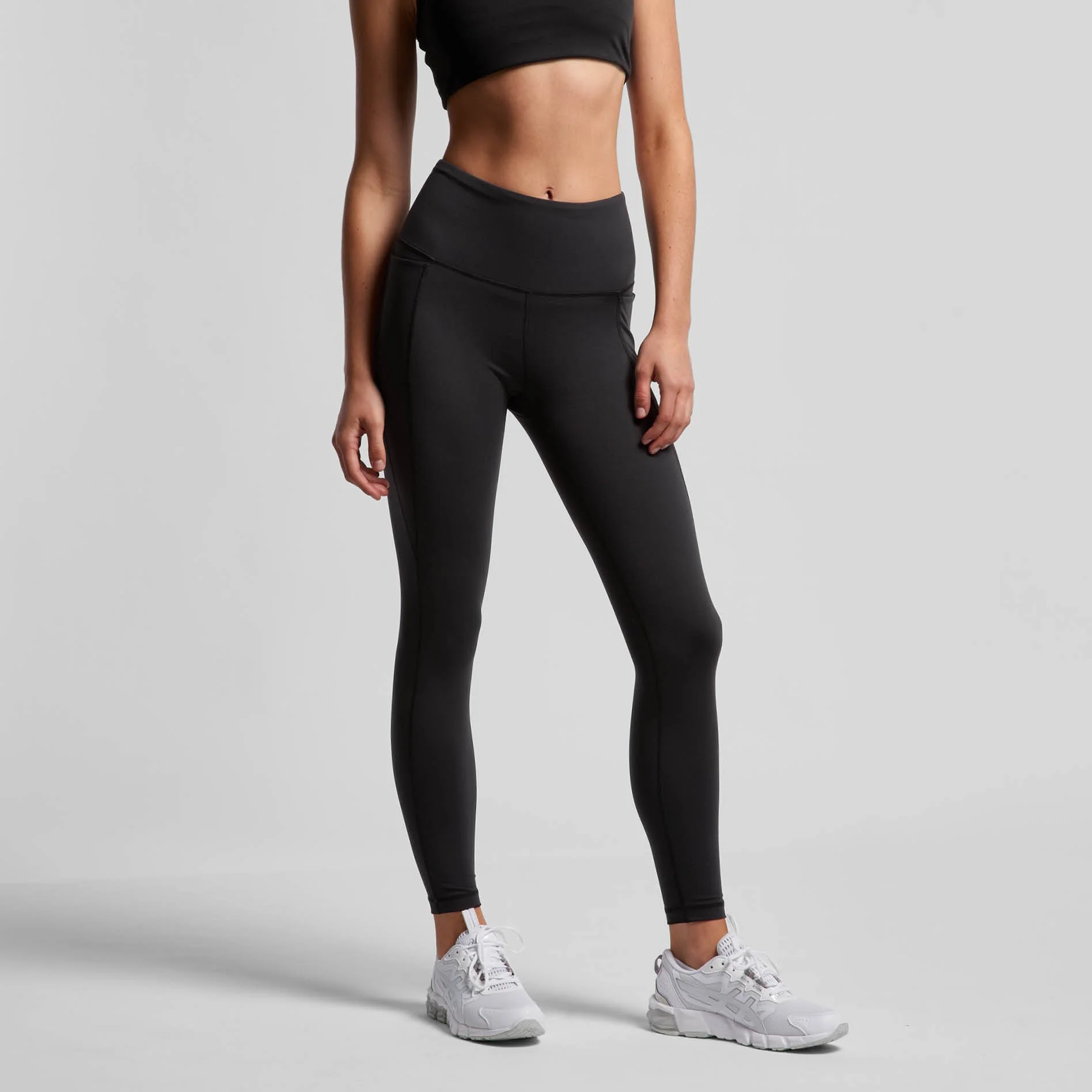 A woman is standing against a plain gray background, wearing a black crop top, black high-waisted athletic leggings, and white sneakers. Her left hand hangs down by her side, and her right hand is partially visible. The image focuses on her midsectio