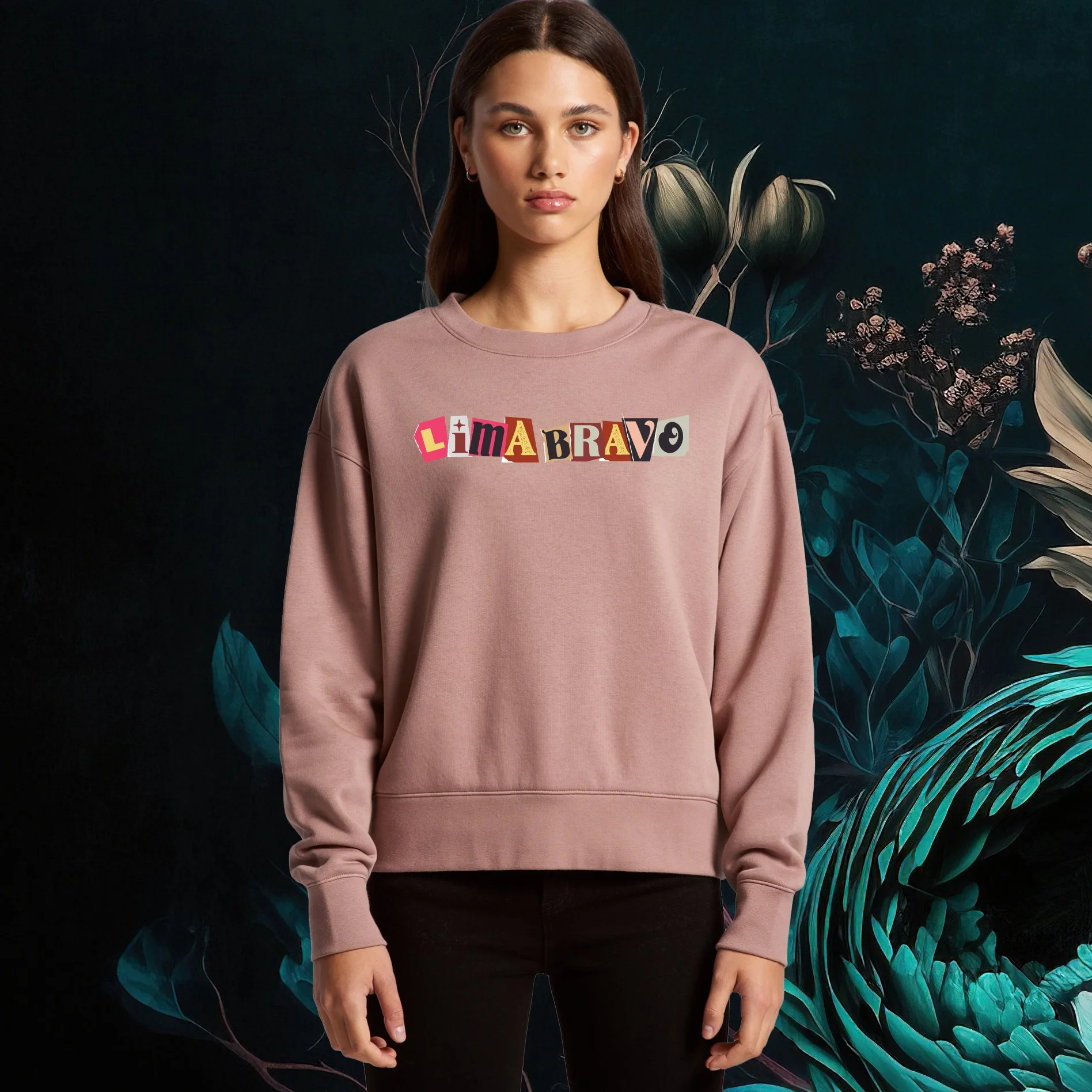 A woman wearing a pink sweatshirt with colorful cut-out letters spelling 'LIMA BRAVO' on the front, standing against a dark background with large leaves and flowers.
