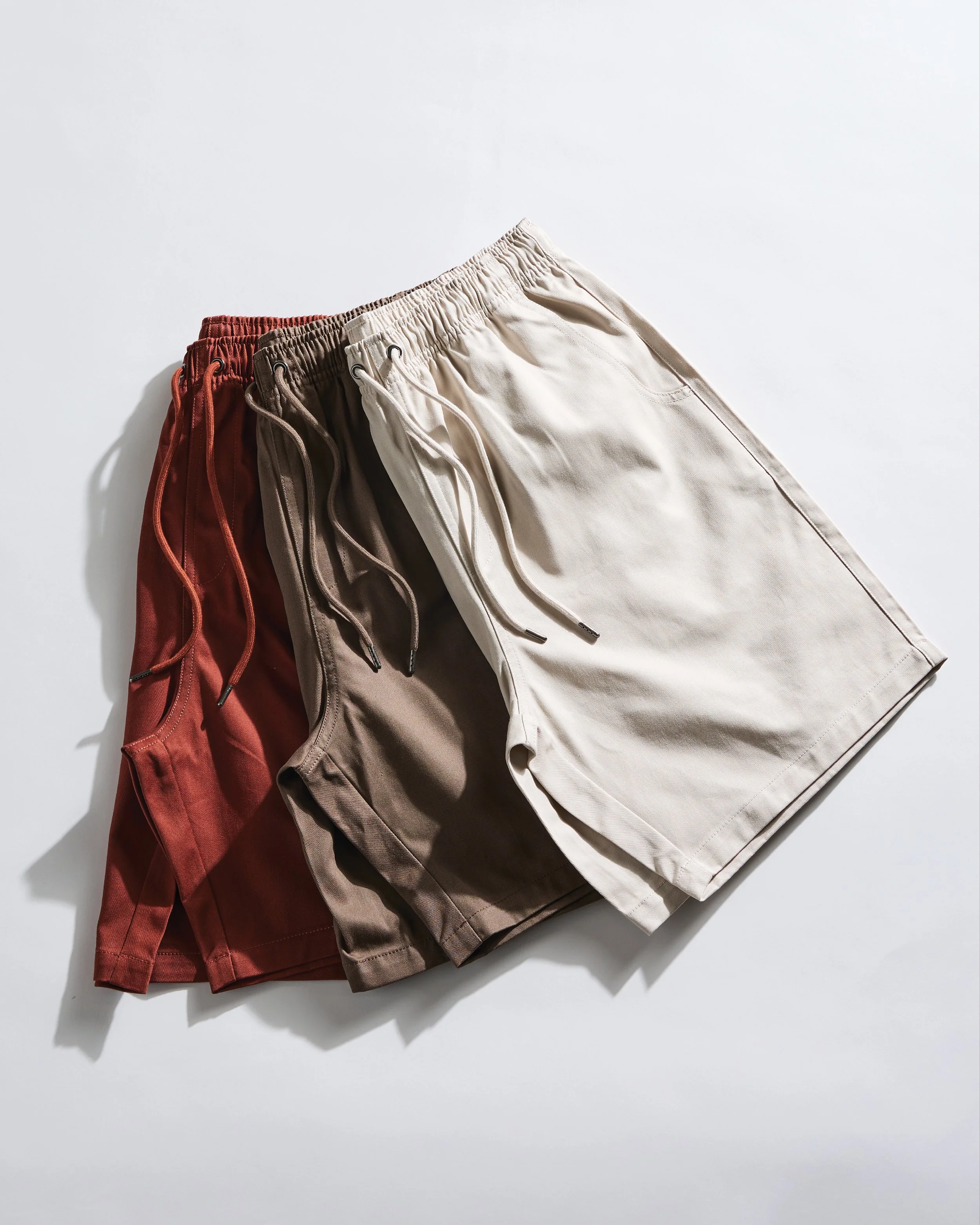 Three pairs of men's shorts in red, beige, and gray colors, laid out on a white surface with shadows.