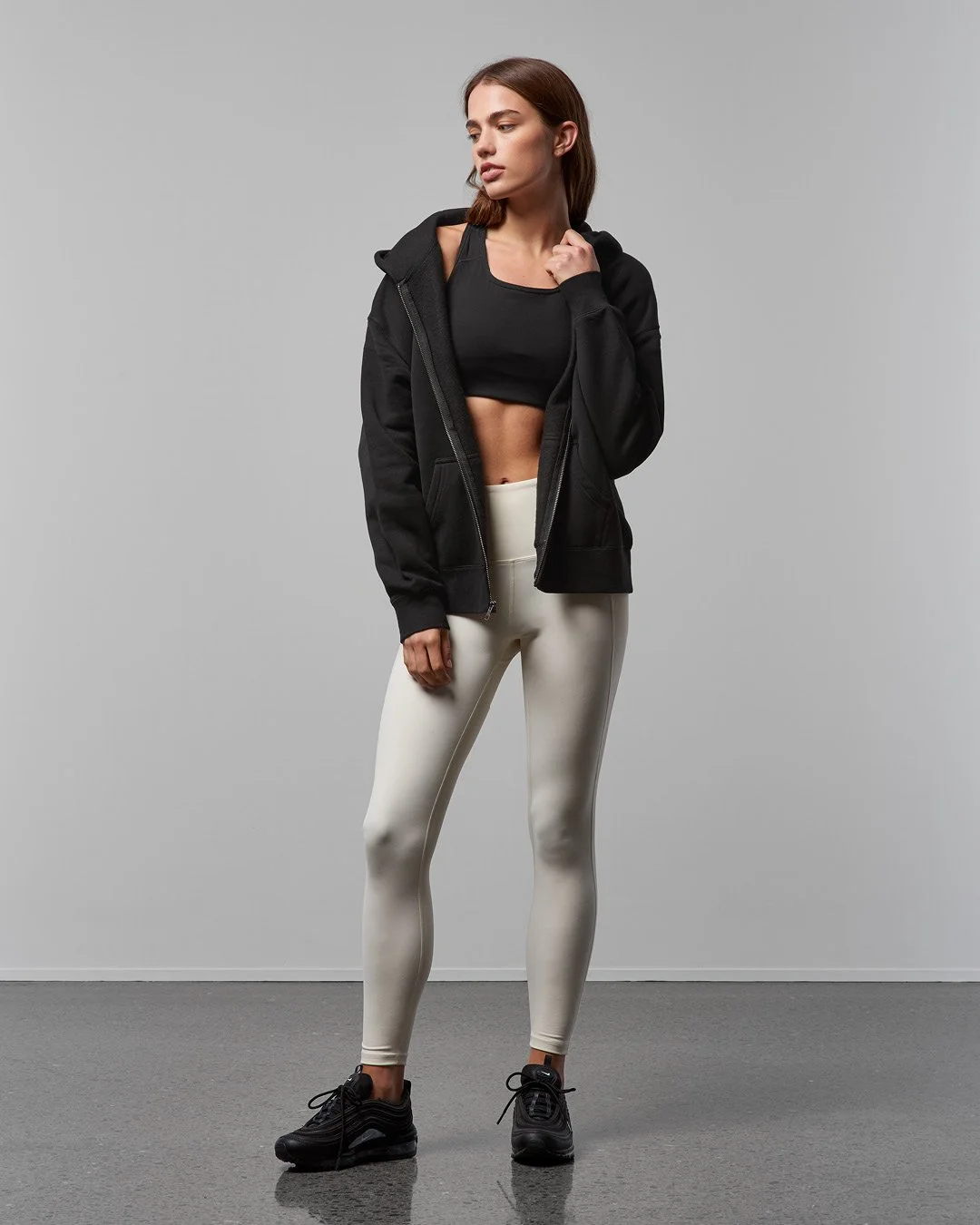 A young woman with brown hair wearing a black cropped top, black zip-up hoodie, white leggings, and black athletic shoes, standing against a plain gray background.