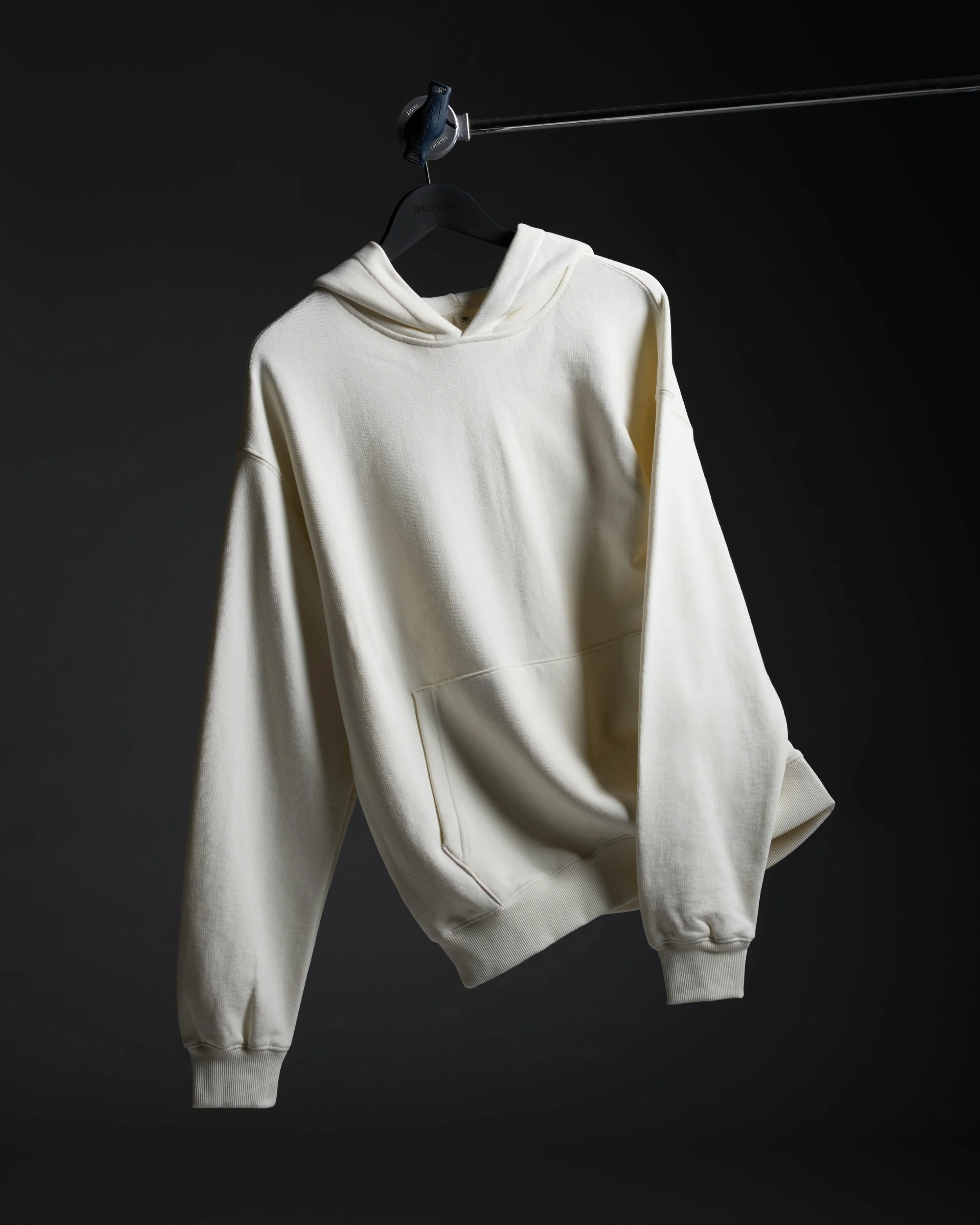 A cream-colored hoodie hangs on a black hanger from a metal rod against a dark background.