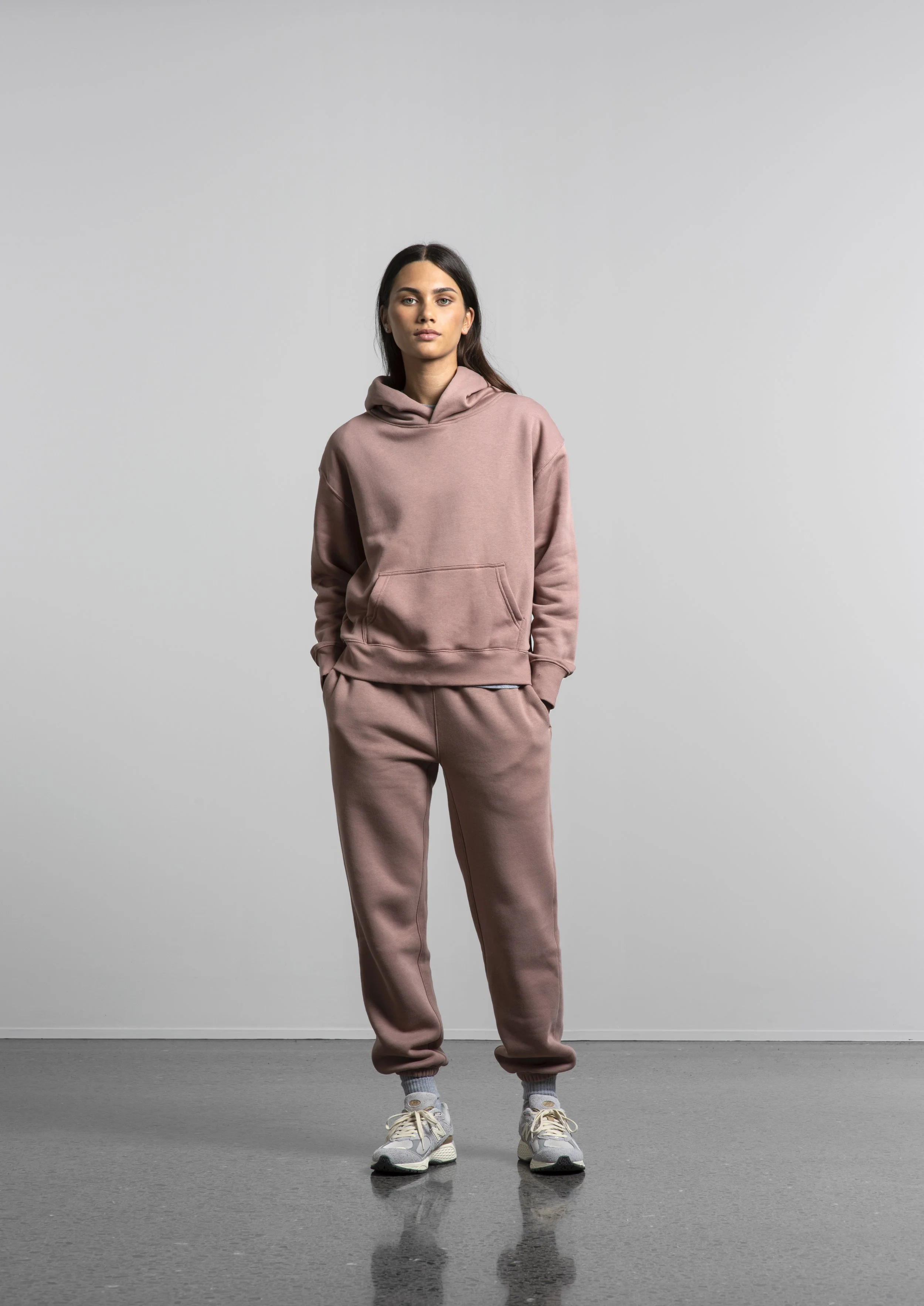 A woman in a pink hoodie and matching sweatpants stands with her hands in her pockets, wearing sneakers, against a plain gray background.