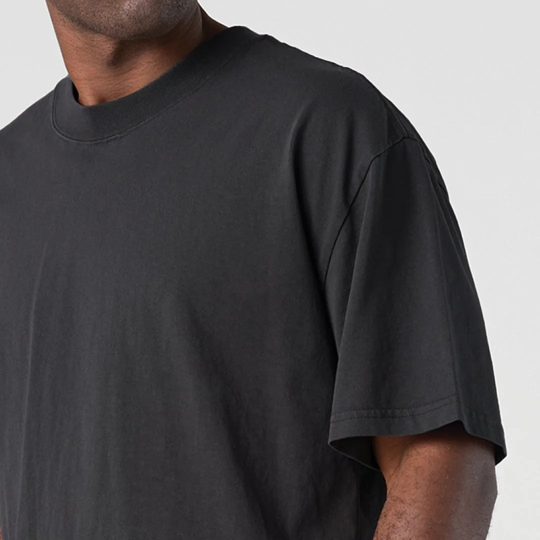 Close-up of a man wearing a plain black t-shirt with a round neckline, showing the shoulder and part of the arm against a plain light gray background.