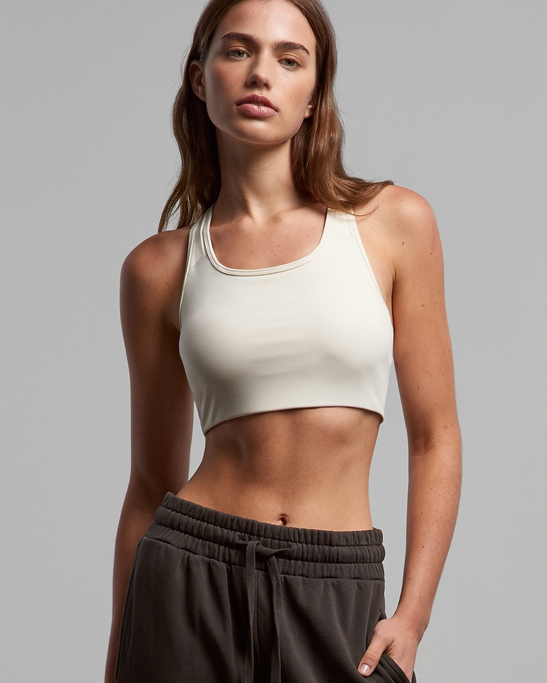 Young woman with red hair wearing a white sports bra and gray sweatpants against a plain background.