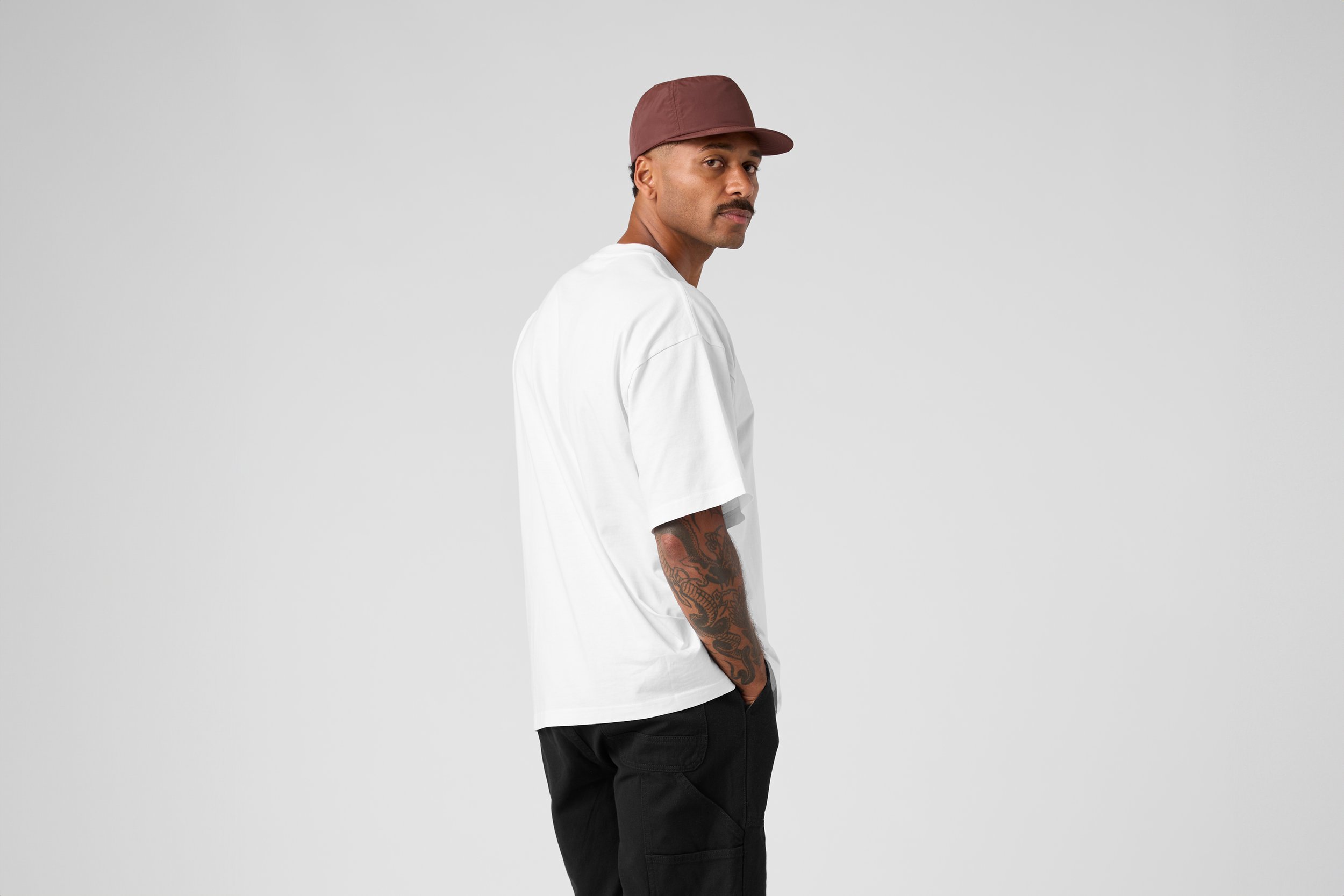 A man with a mustache wearing a maroon cap, white t-shirt, and black pants, standing against a plain light gray background.