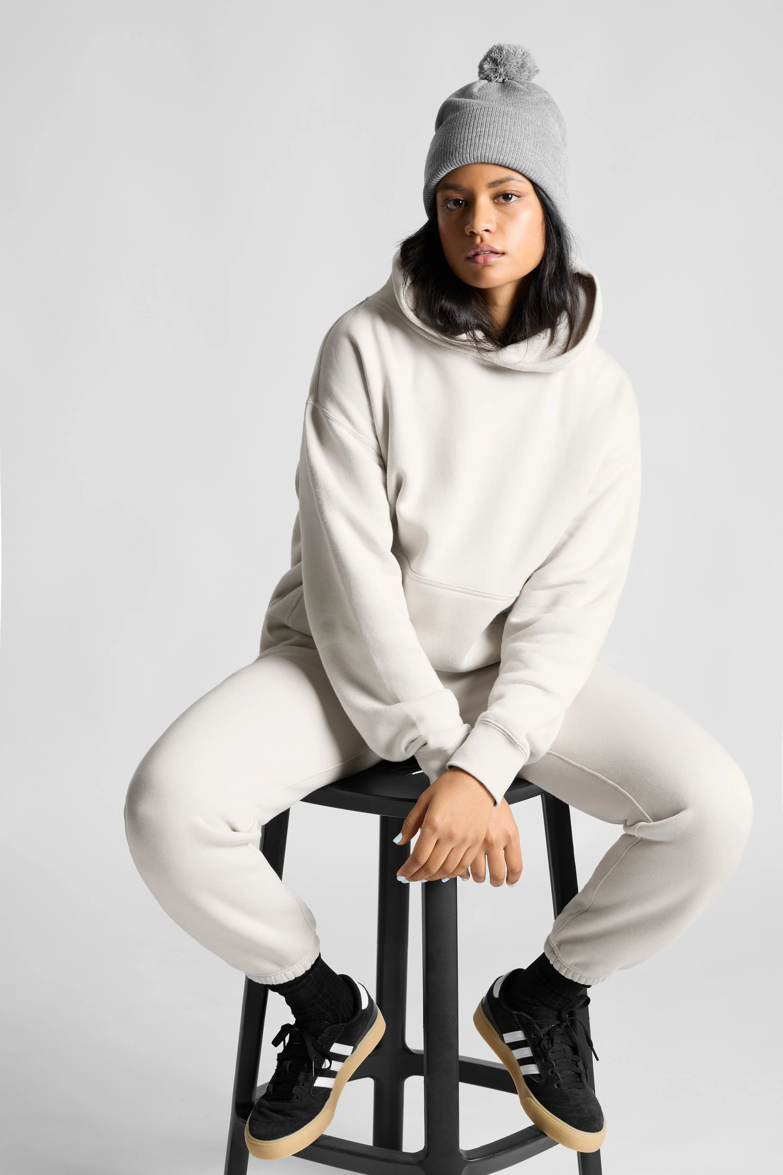A young woman with medium skin tone, black hair, and wearing a gray knit beanie with a pom-pom, sitting on a black stool. She has on a cream hoodie and matching sweatpants, and wears black sneakers with white stripes and gum soles. The background is 
