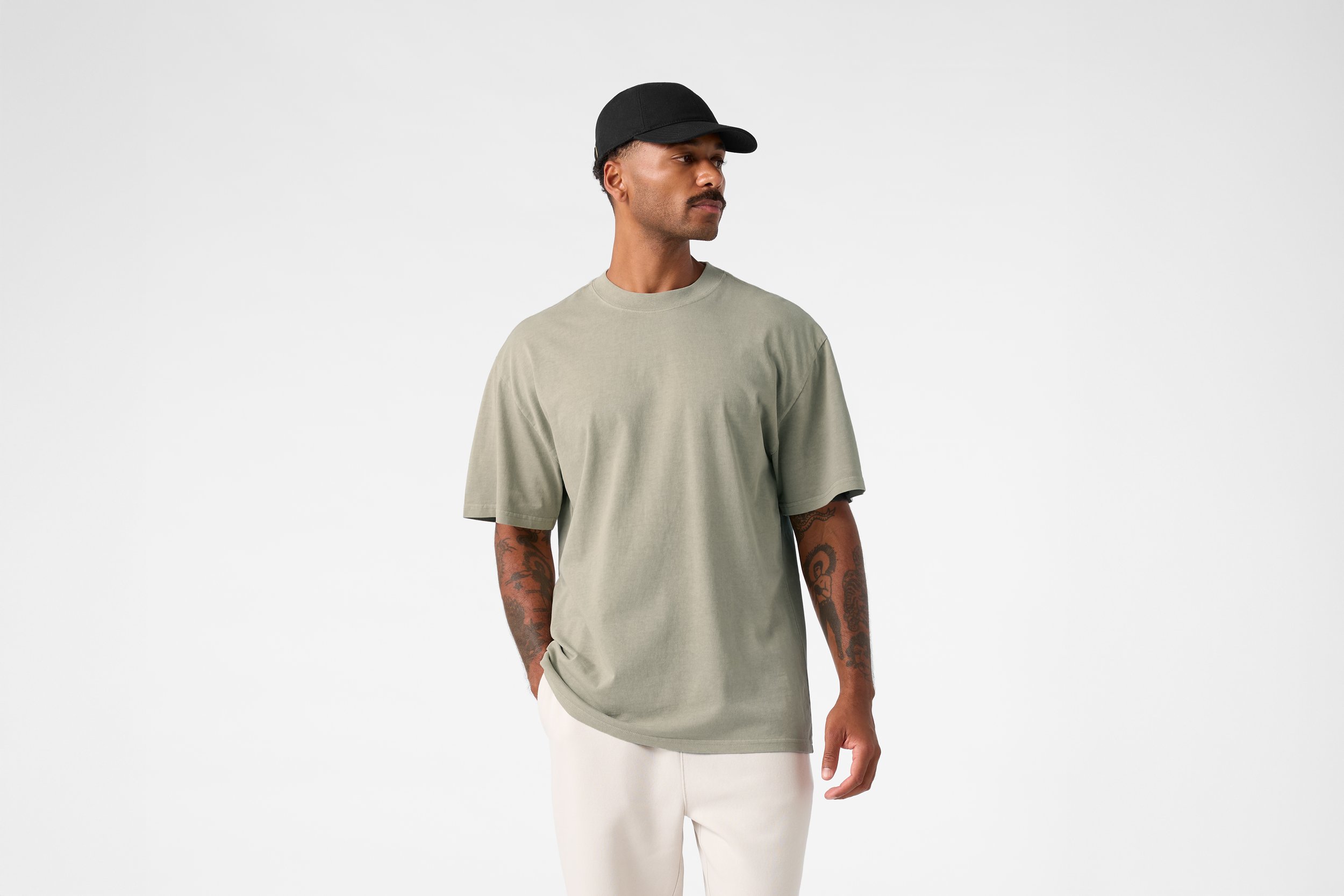 A man wearing a black cap, a loose light green T-shirt, and light-colored pants, standing with one hand in his pocket against a plain white background.
