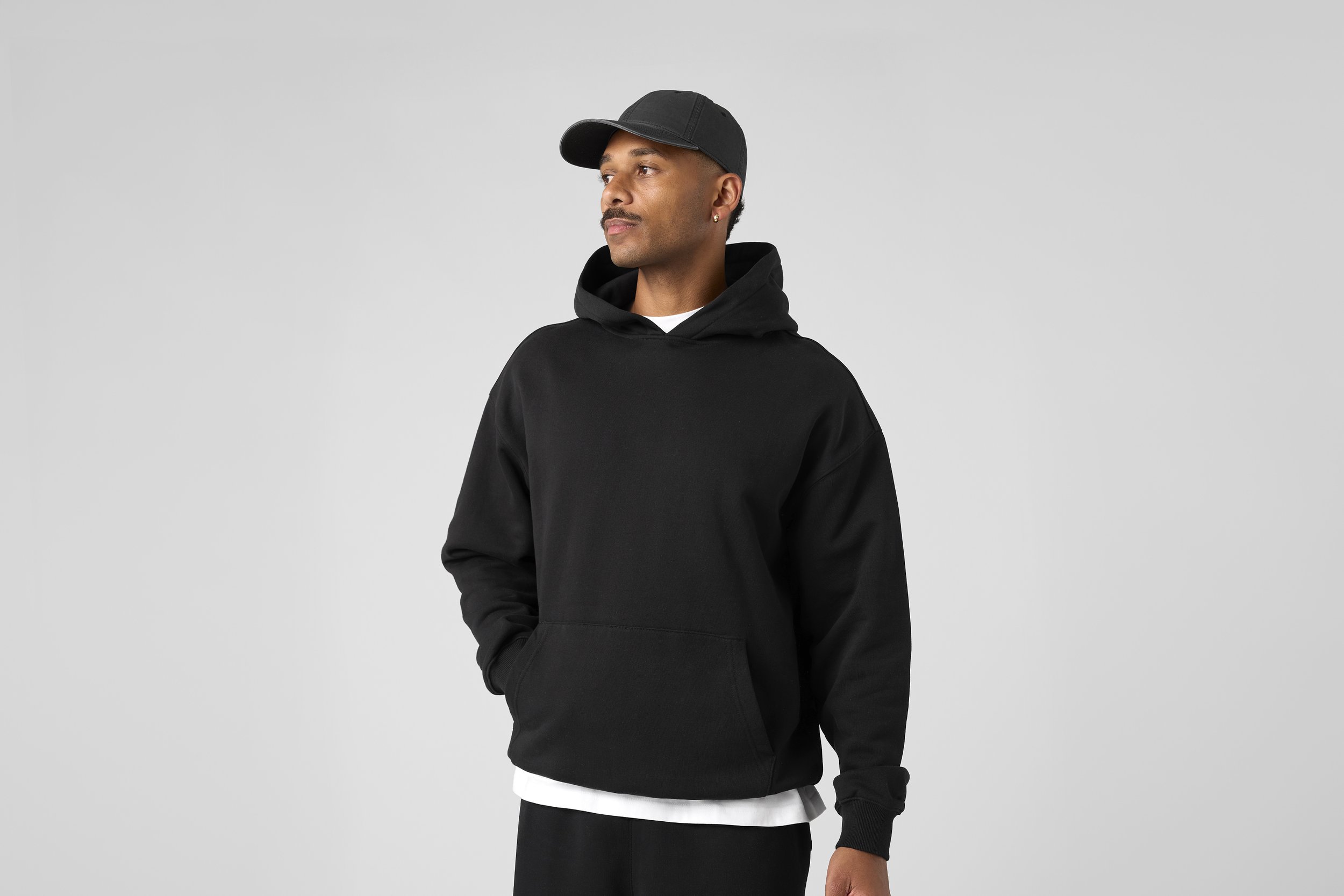 A man wearing a black baseball cap, black hoodie, and black pants standing against a plain light gray background.