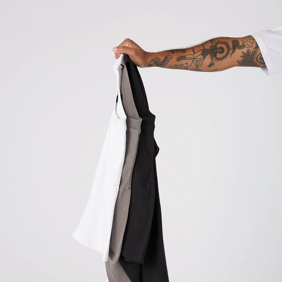 Person with tattooed arm holding a hanger with three pairs of pants against a plain background.