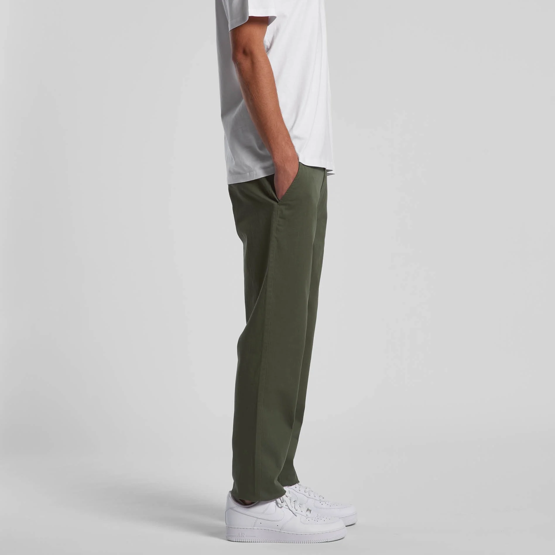 Side view of a person wearing white sneakers, dark green pants, white T-shirt, and standing with their hand in their pocket against a plain light gray background.