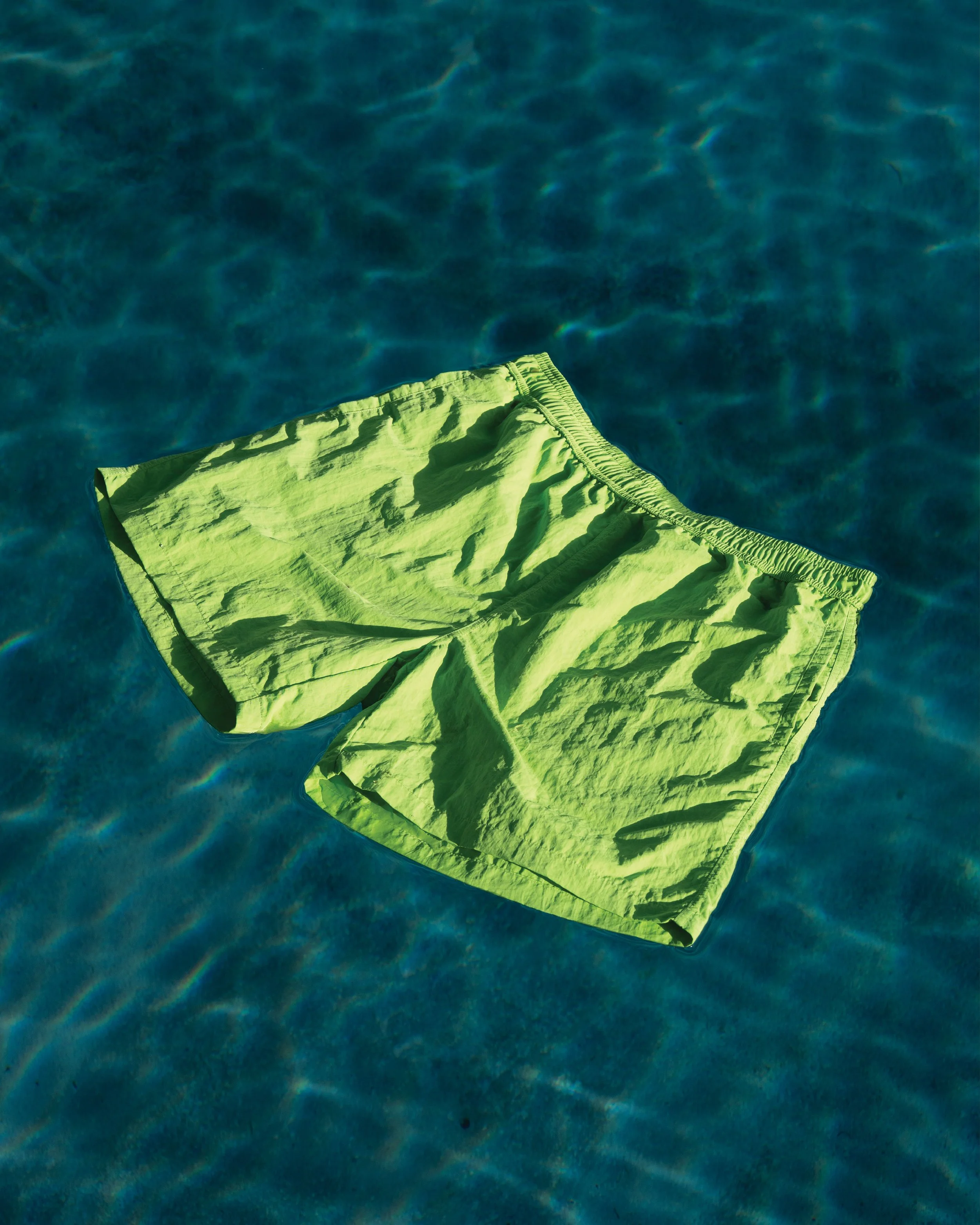 Bright green swim shorts floating on a clear blue swimming pool with sunlight reflections.