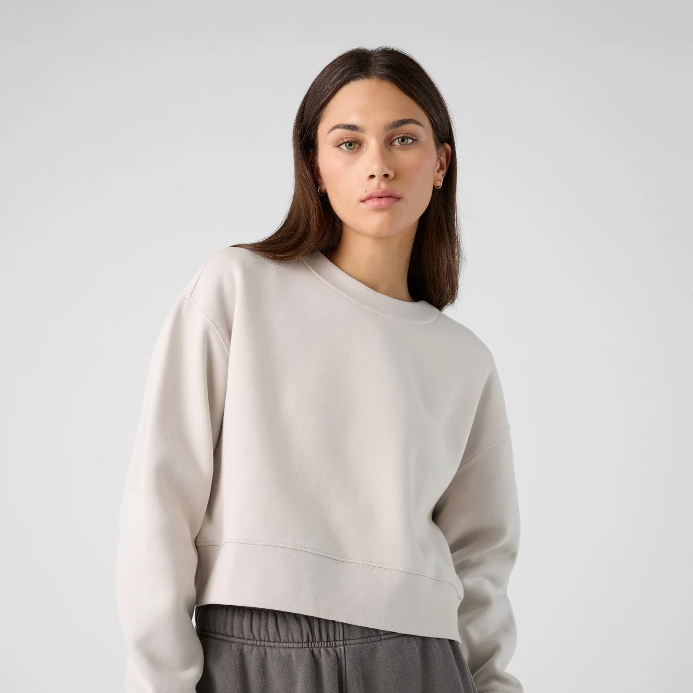 A young woman with straight brown hair, wearing a beige sweatshirt and gray pants, posing against a plain light gray background.